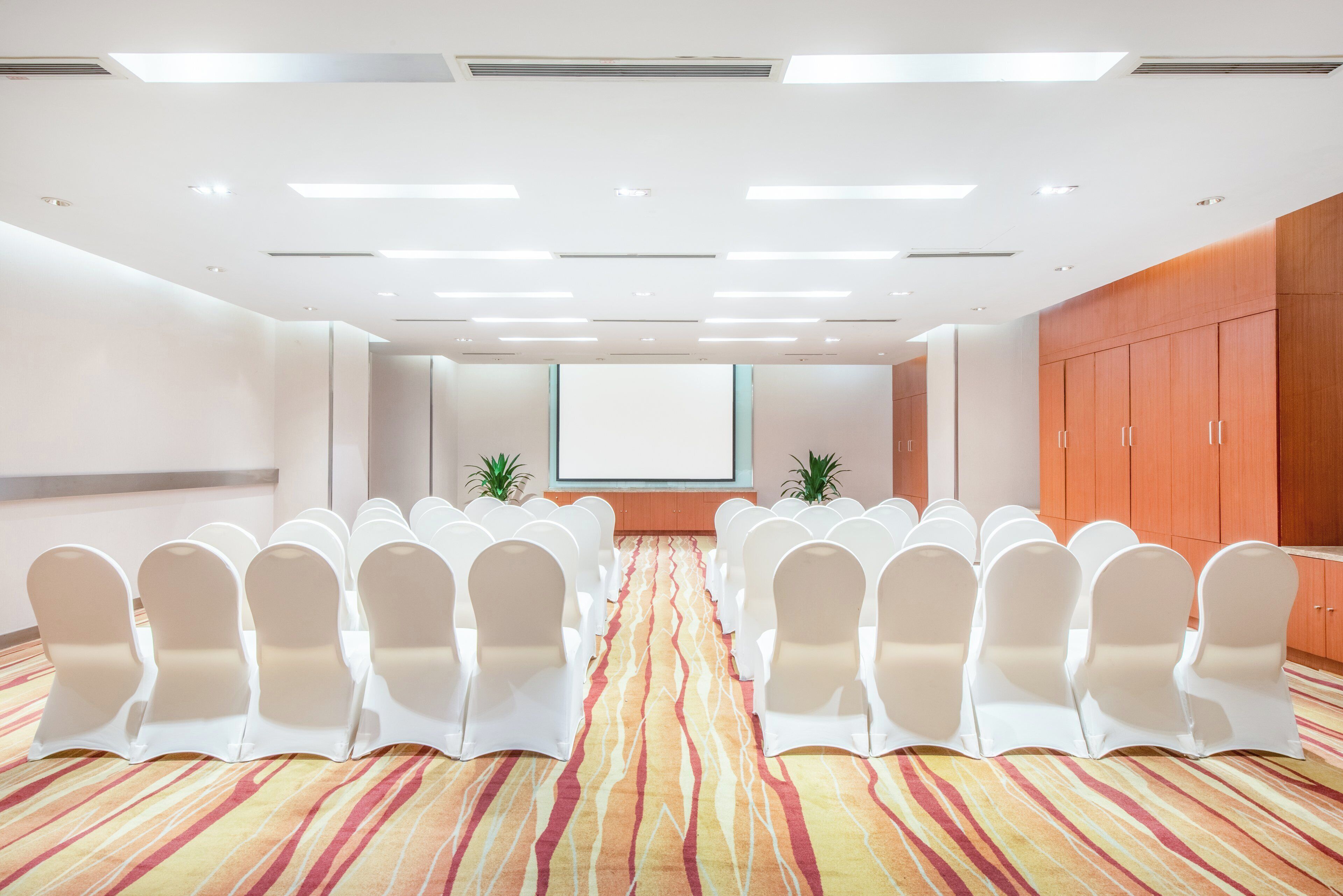 A conference room with rows of white chairs, a projector screen, and a patterned carpet.