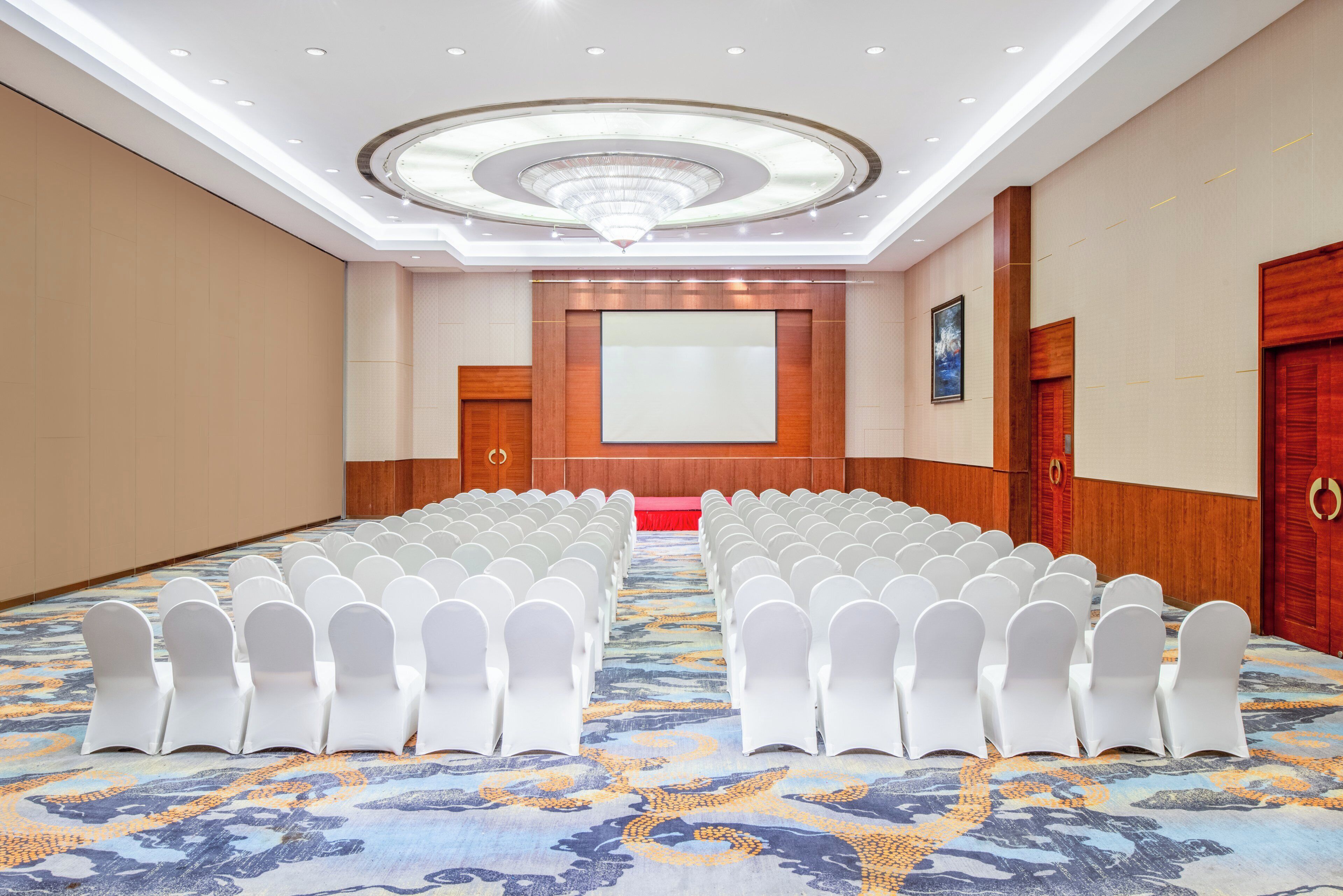 A conference room with rows of white chairs, a large screen, and a patterned carpet.