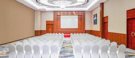 Un salón de conferencias con filas de sillas blancas, una pantalla grande y una alfombra estampada.