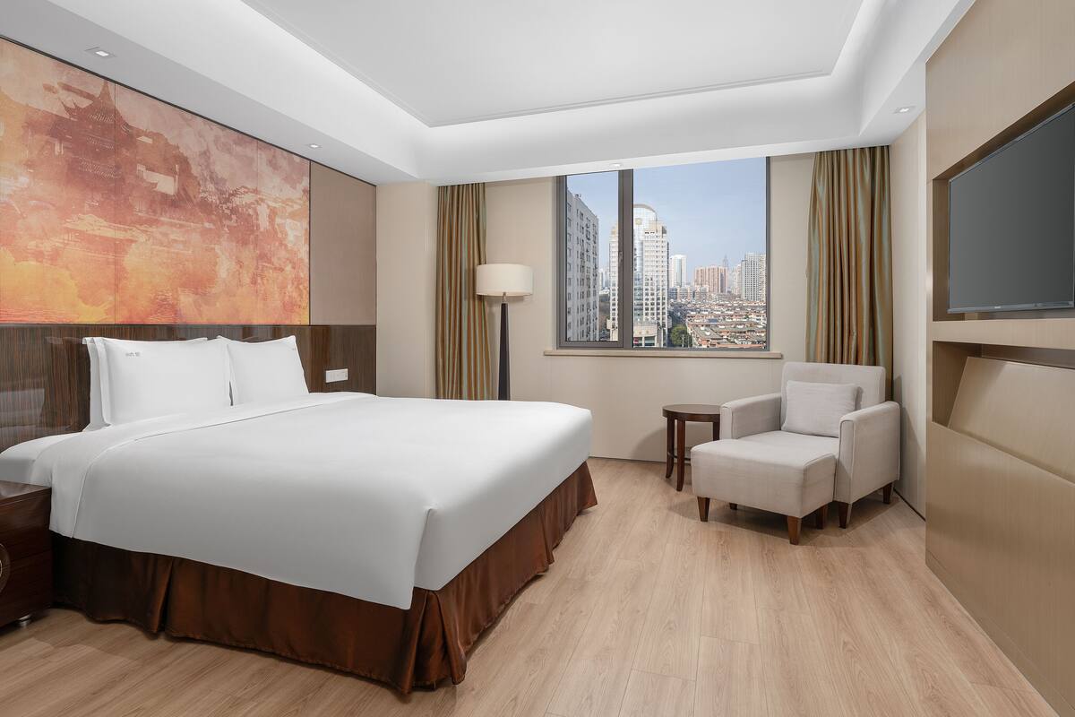 A hotel room with a large bed, a chair, a TV, a window with a city view, and a wall with a large abstract painting.
