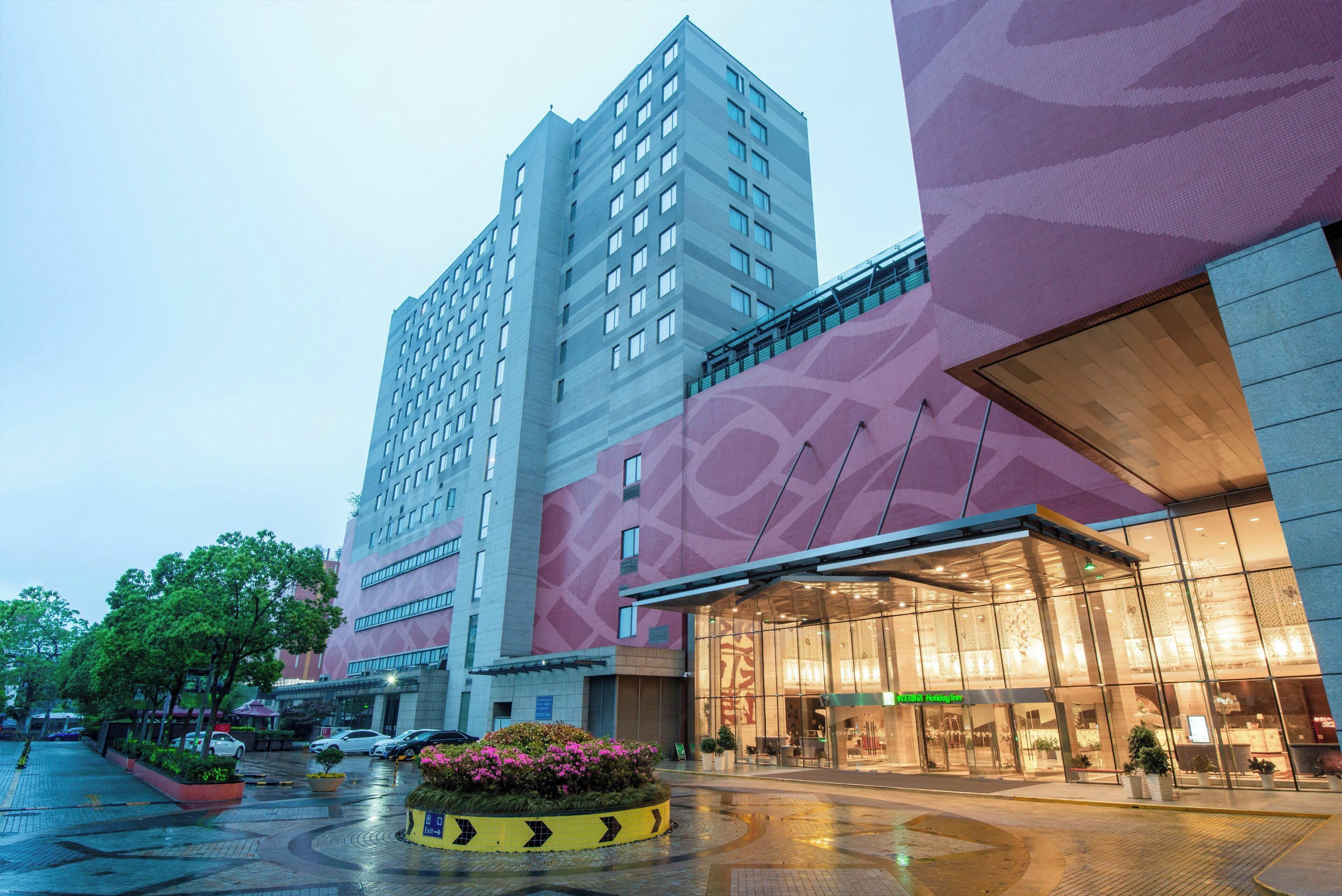 A modern hotel with a prominent glass facade, a central courtyard with a yellow directional sign, and a high-rise building in the background.
