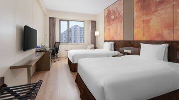 Habitación de hotel con dos camas, un escritorio con televisor, una silla, una lámpara y ventana con vista a la ciudad.