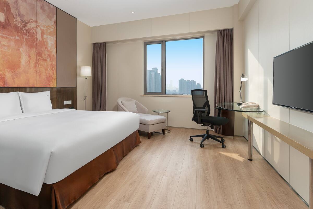 A hotel room with a large bed, a desk with a chair, a flat-screen TV, and a window with a city view.