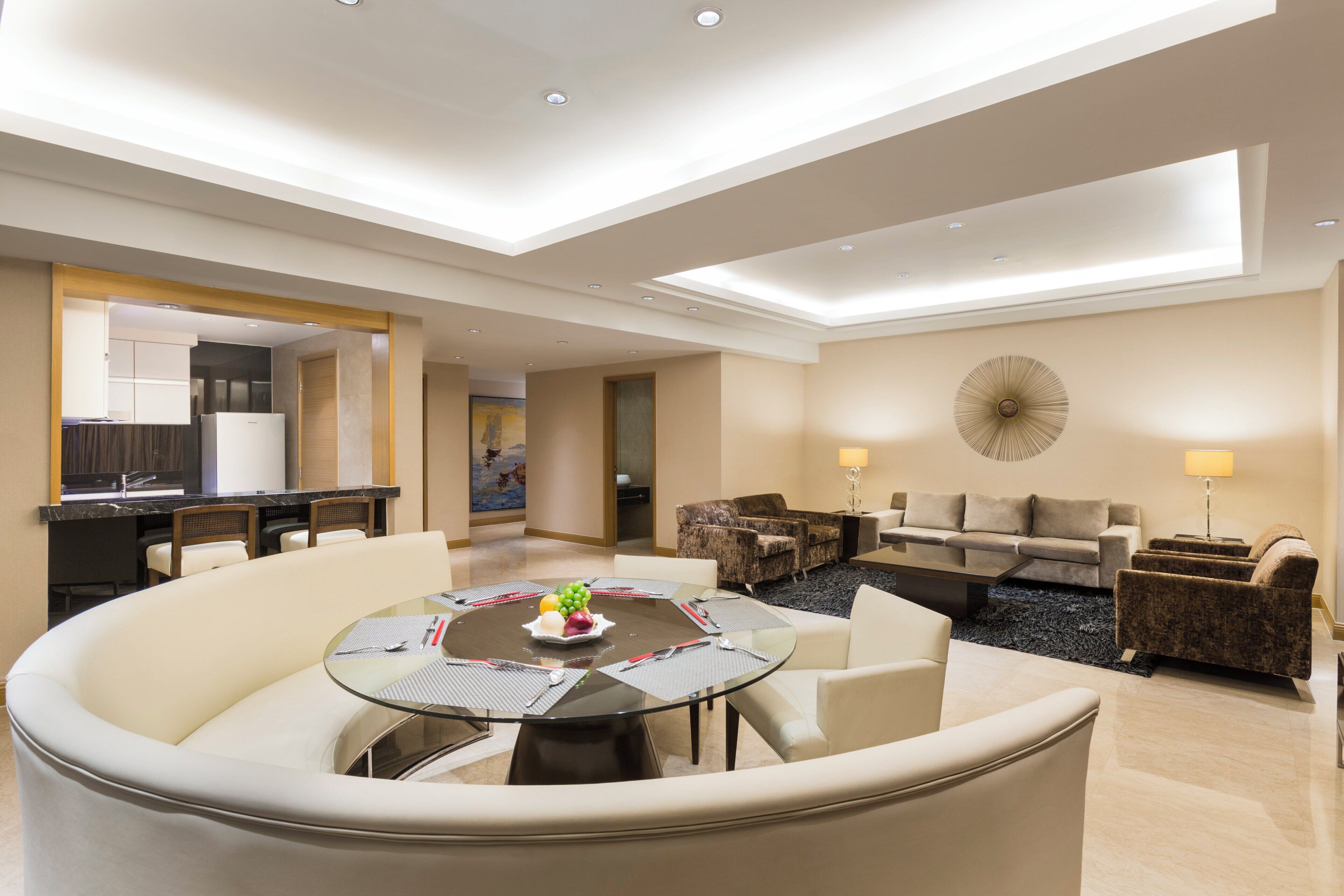 A modern hotel room with a dining area, a living room with a sofa and armchairs, and a kitchen in the background.