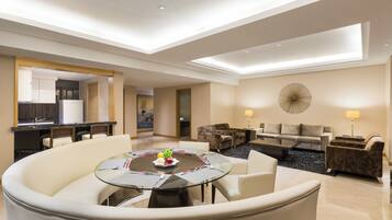 A modern hotel room with a dining area, a living room with a sofa and armchairs, and a kitchen in the background.