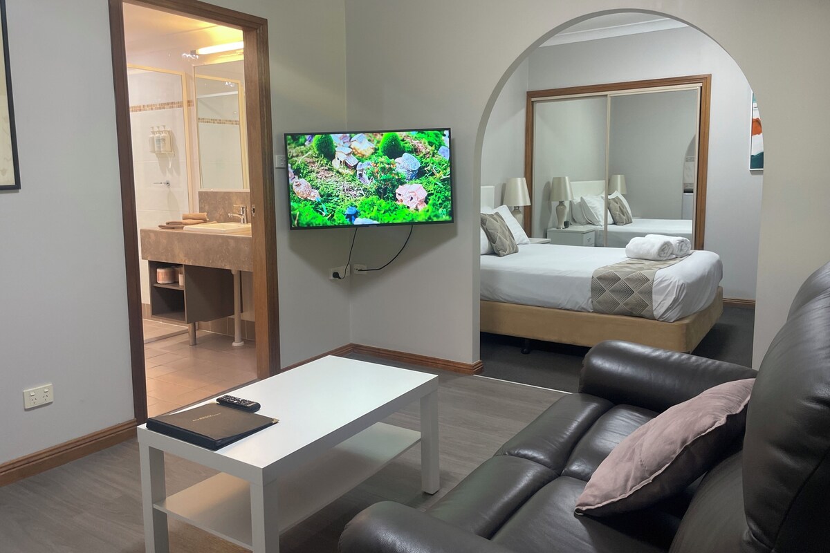 A modern hotel room with a television, a sofa, a bed, and a bathroom visible through an archway.