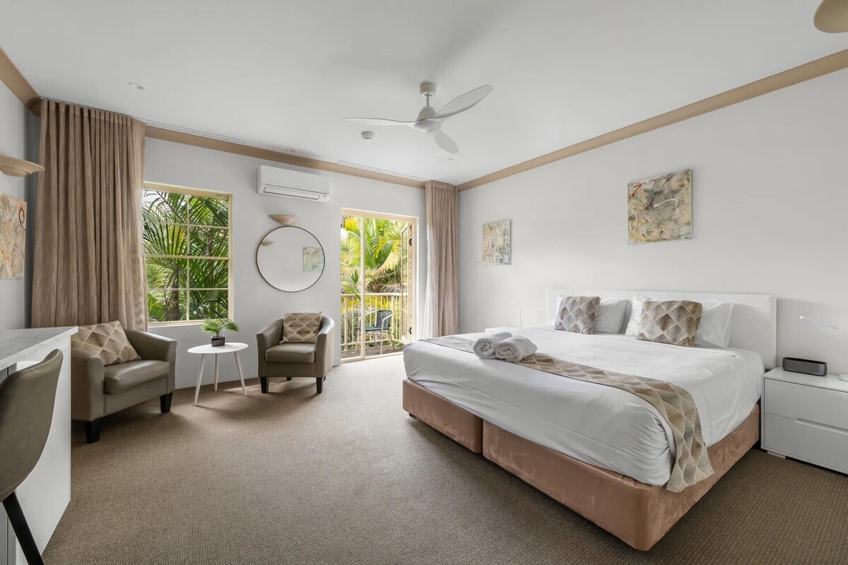 A hotel room with a bed, a desk, a chair, a small table, and a ceiling fan.