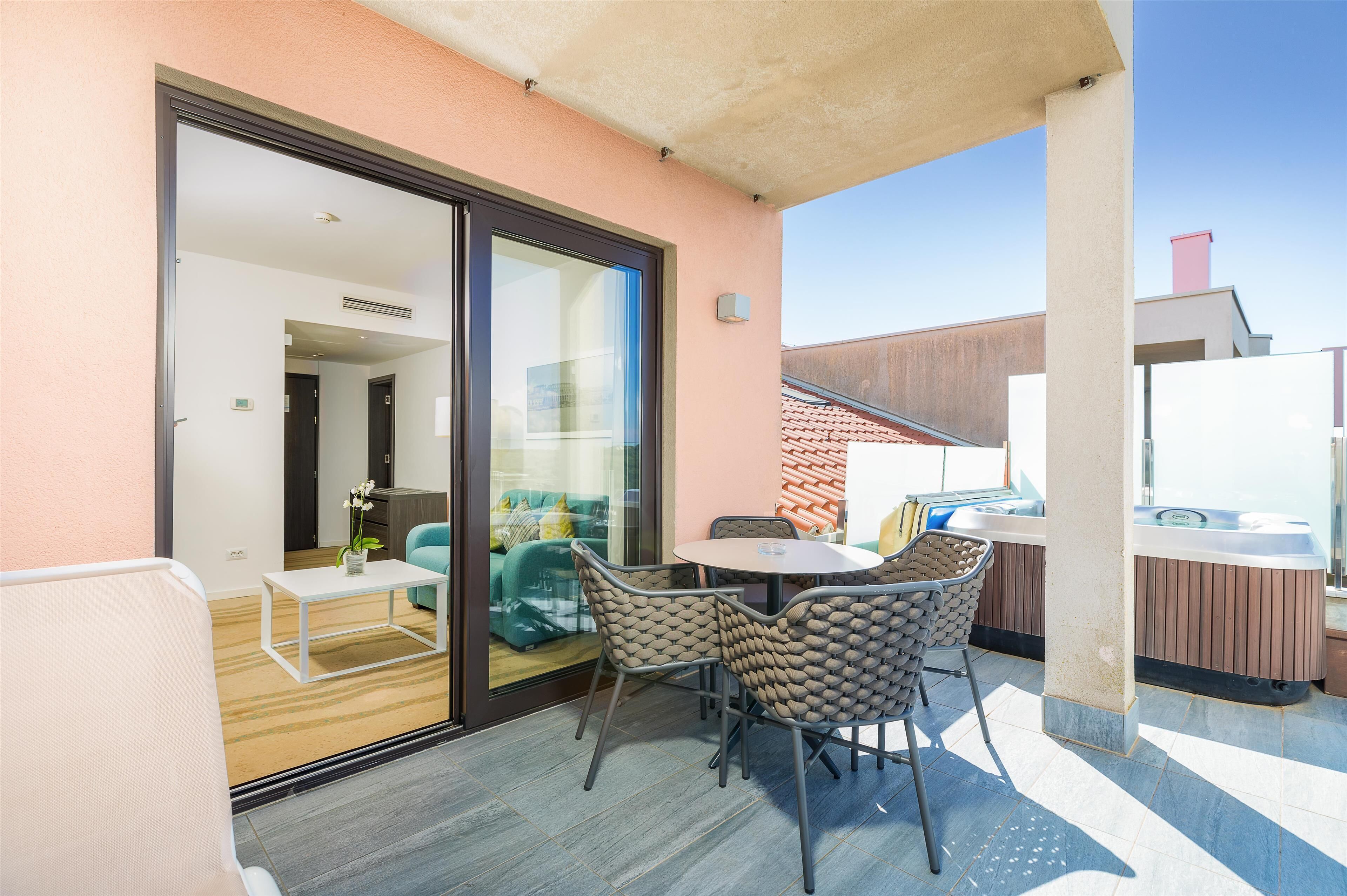 A balcony with a glass door leading to a room, a round table with chairs, and a hot tub.