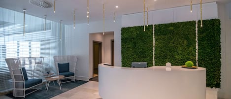 A reception area with a white reception desk, green wall, hanging lights, and seating area with chairs and a table.
