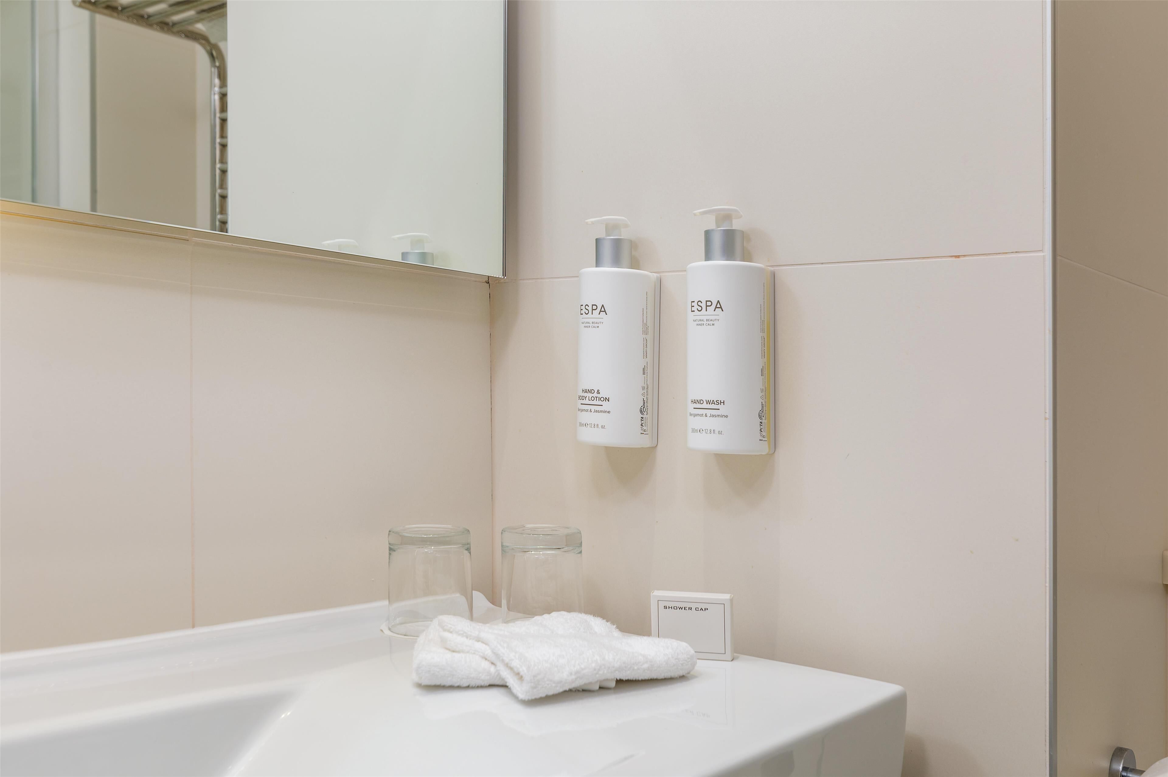 Une salle de bain avec un lavabo blanc, deux flacons de savon pour les mains de la marque Espa, une serviette et un bonnet de douche posés sur le comptoir.