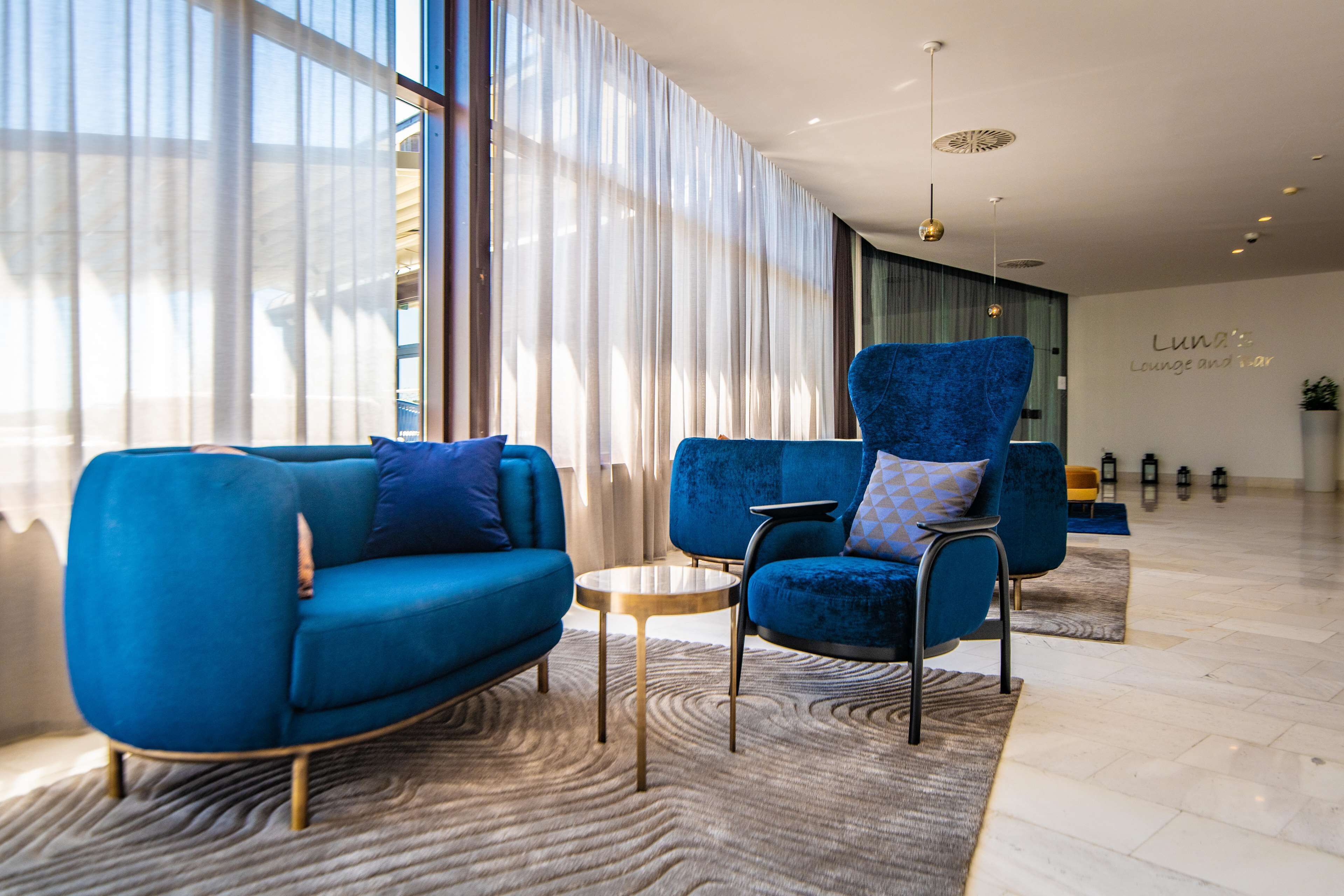 A modern lounge area with blue sofas, a round coffee table, and a large window with sheer curtains.