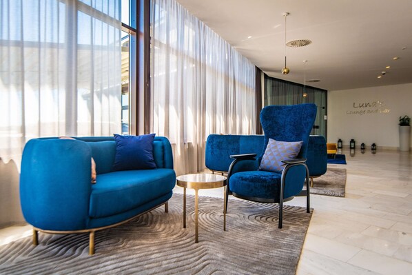 A modern lounge area with blue sofas, a round coffee table, and a large window with sheer curtains.
