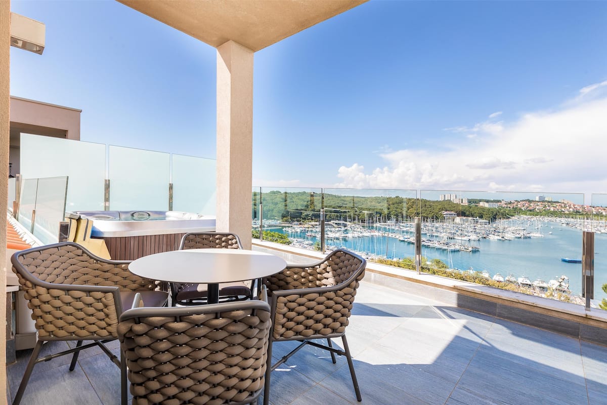 A terrace with a round table and wicker chairs, overlooking a marina with boats and a cityscape.