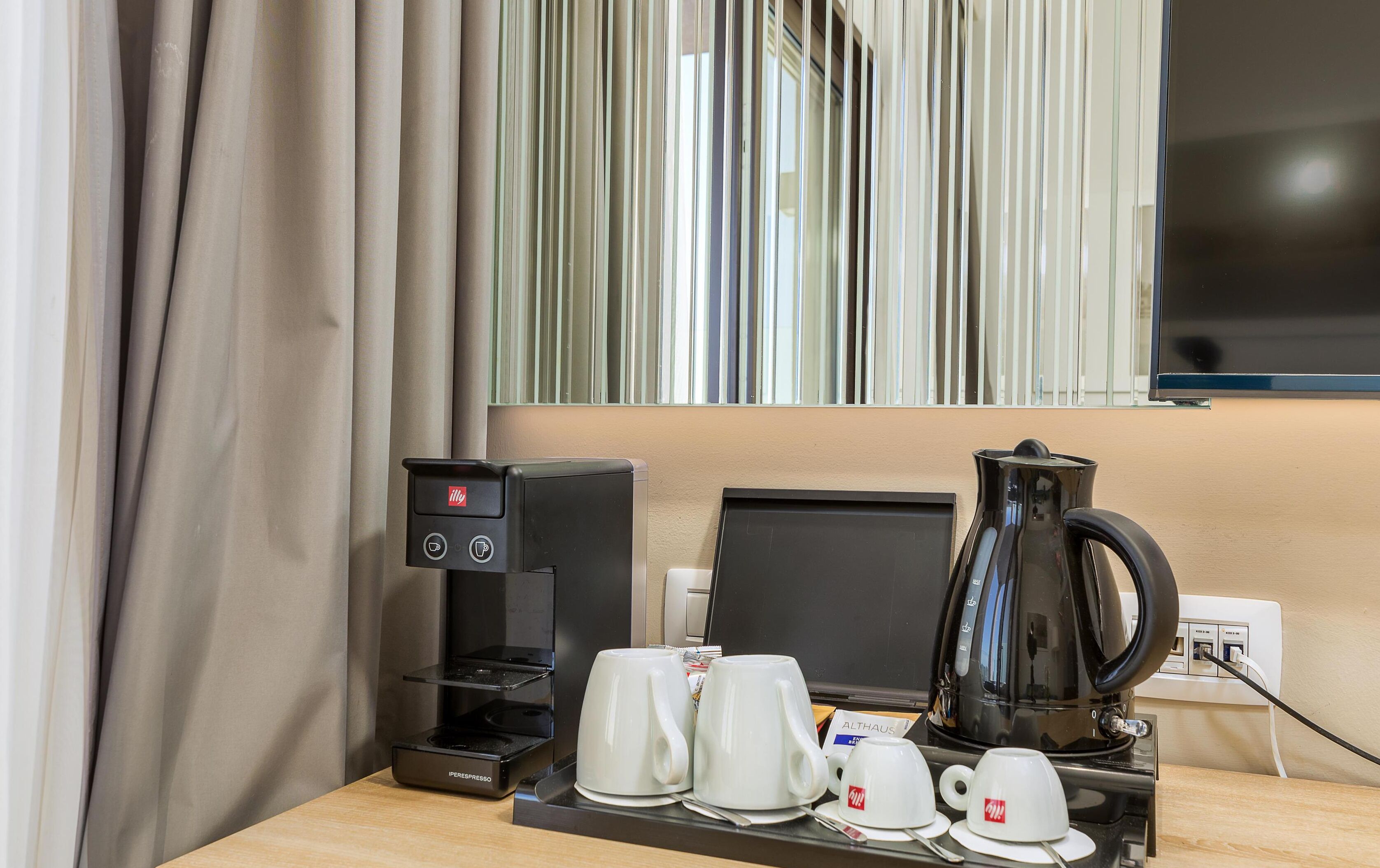 Una habitación de hotel con cafetera, dos tazas blancas y un hervidor sobre una mesa de madera.