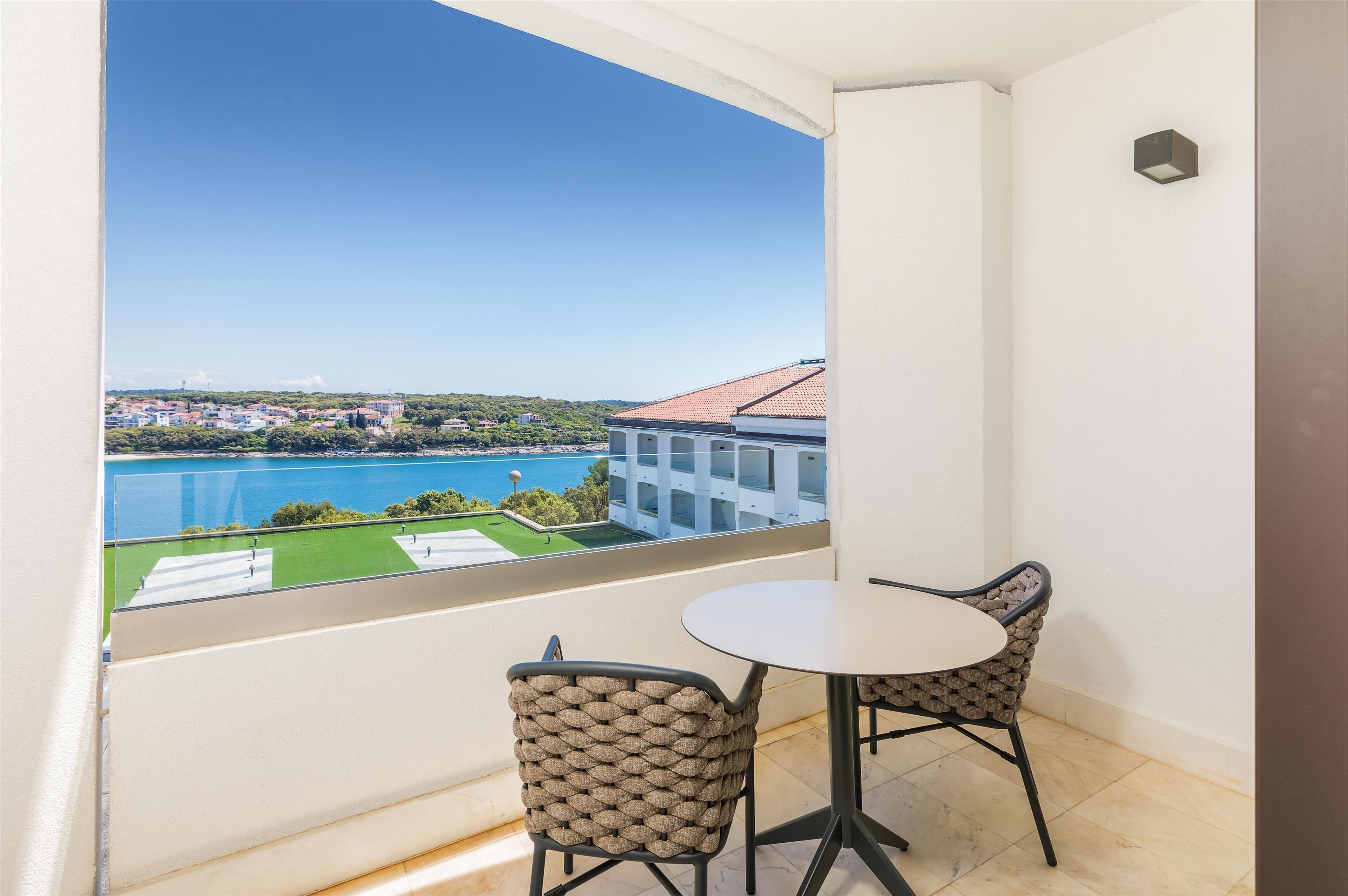 A balcony with a view of a lake and buildings, a round table, and two wicker chairs.
