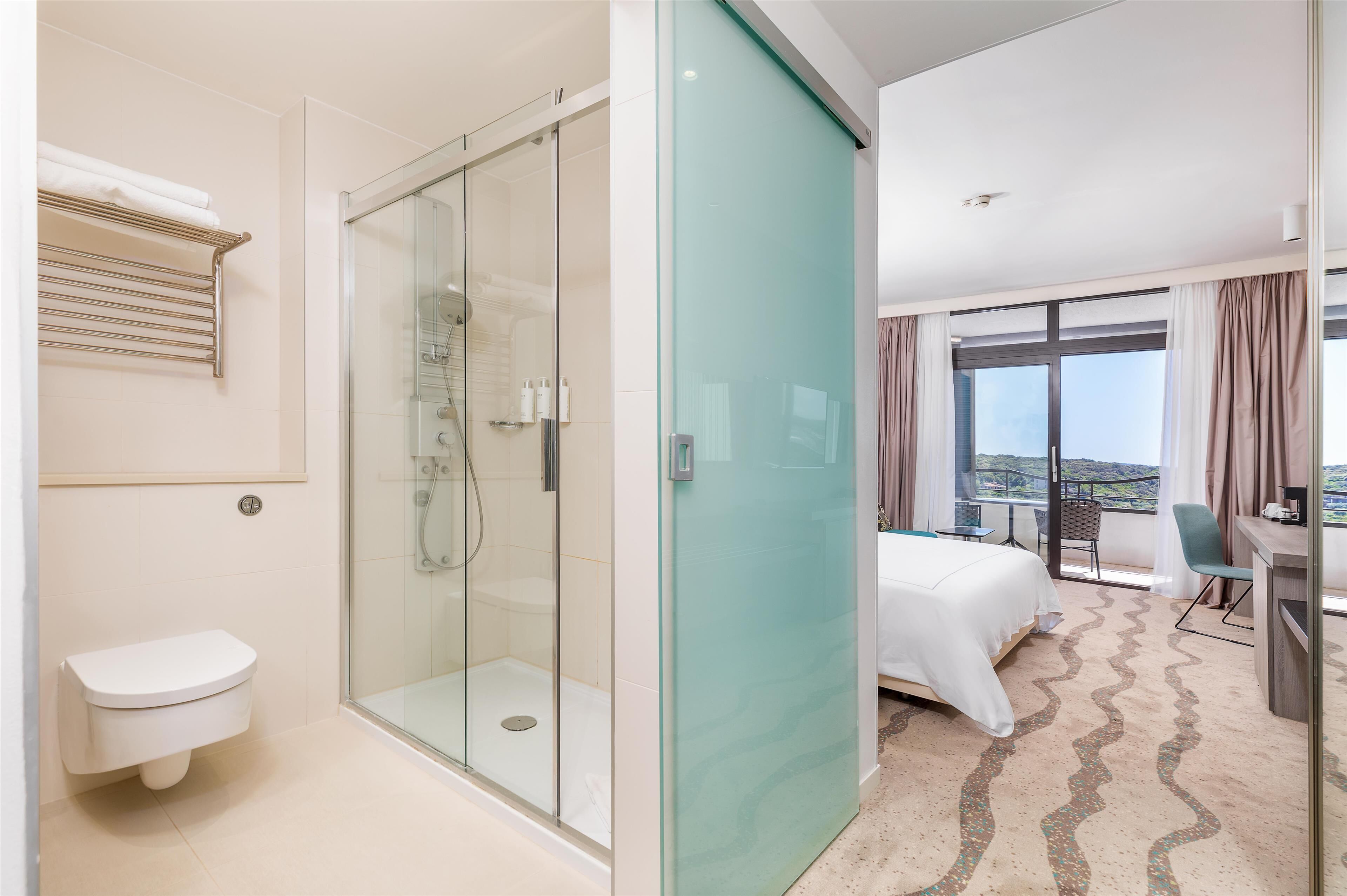 Une chambre d’hôtel moderne dotée d’une douche en verre, de toilettes blanches, d’un lit avec une couette blanche, d’un bureau et offrant une vue sur le paysage grâce à de grandes fenêtres.