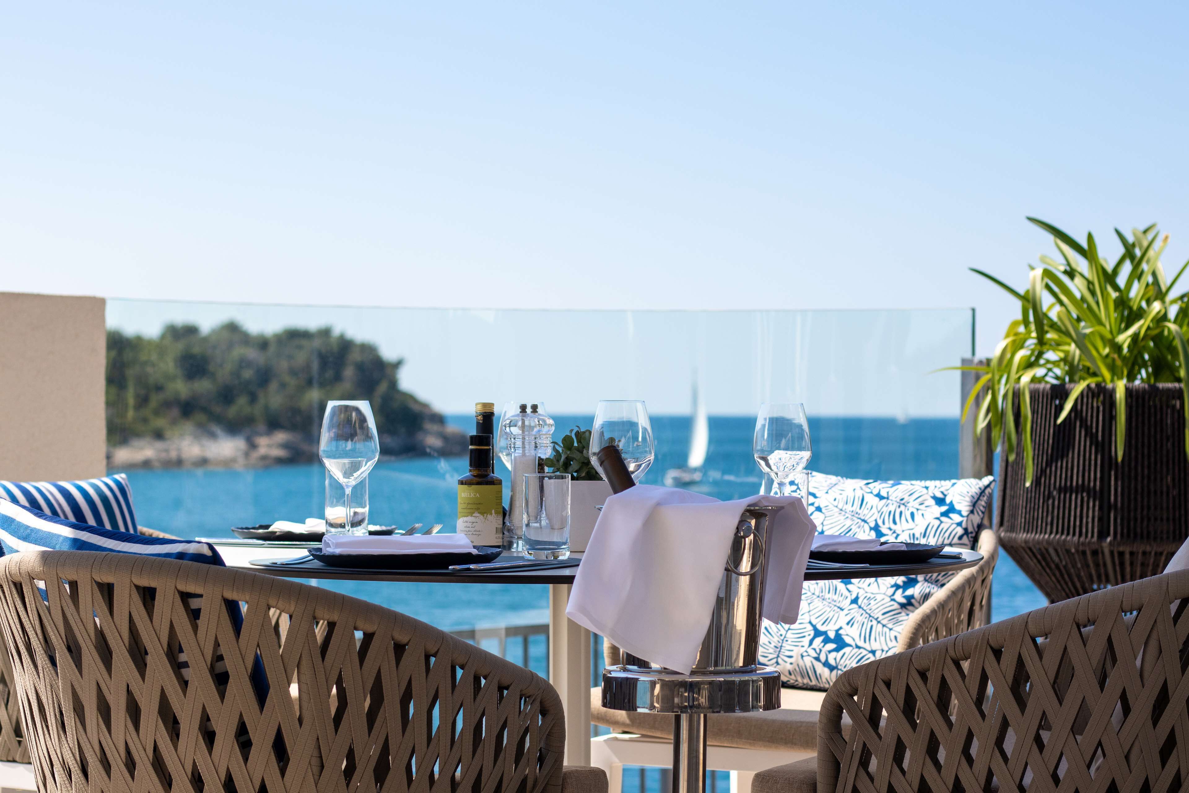 Tavola imbandita con bicchieri da vino, bottiglie e menu, con vista panoramica sulla costa.