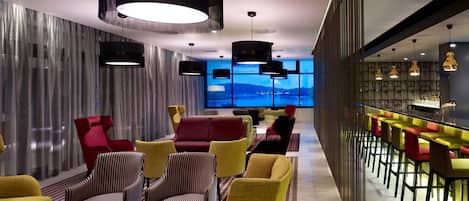 Un área de descanso moderna con un bar, asientos con sillas de colores y vistas a una masa de agua a través de grandes ventanales.