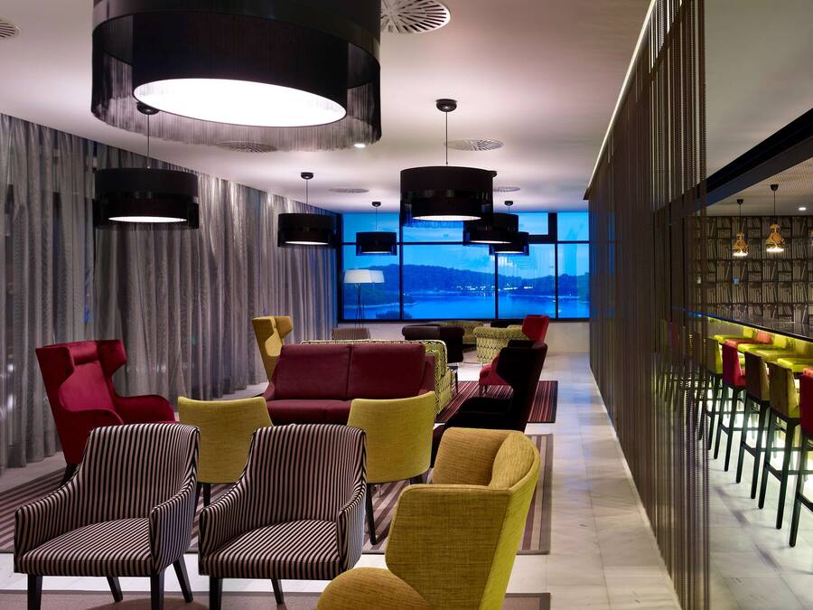 A modern lounge area with a bar, seating with colorful chairs, and a view of a body of water through large windows.
