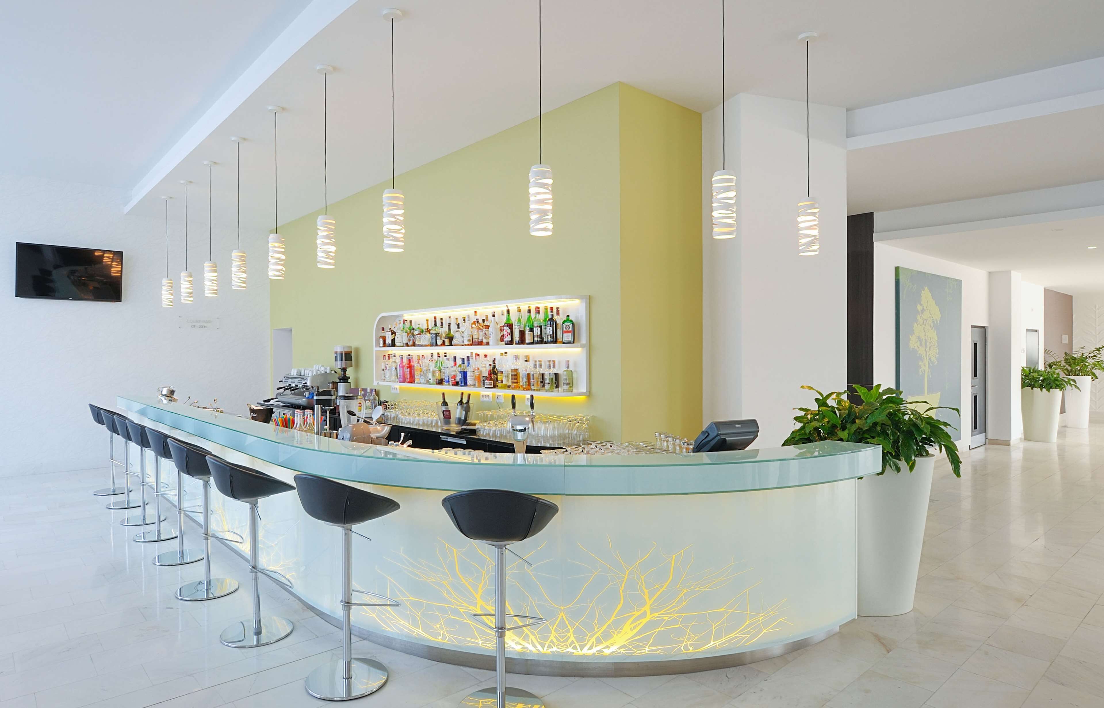 Un espace bar moderne doté d’un comptoir en verre, de plusieurs tabourets de bar, de luminaires suspendus et d’une étagère remplie de bouteilles et de verres.