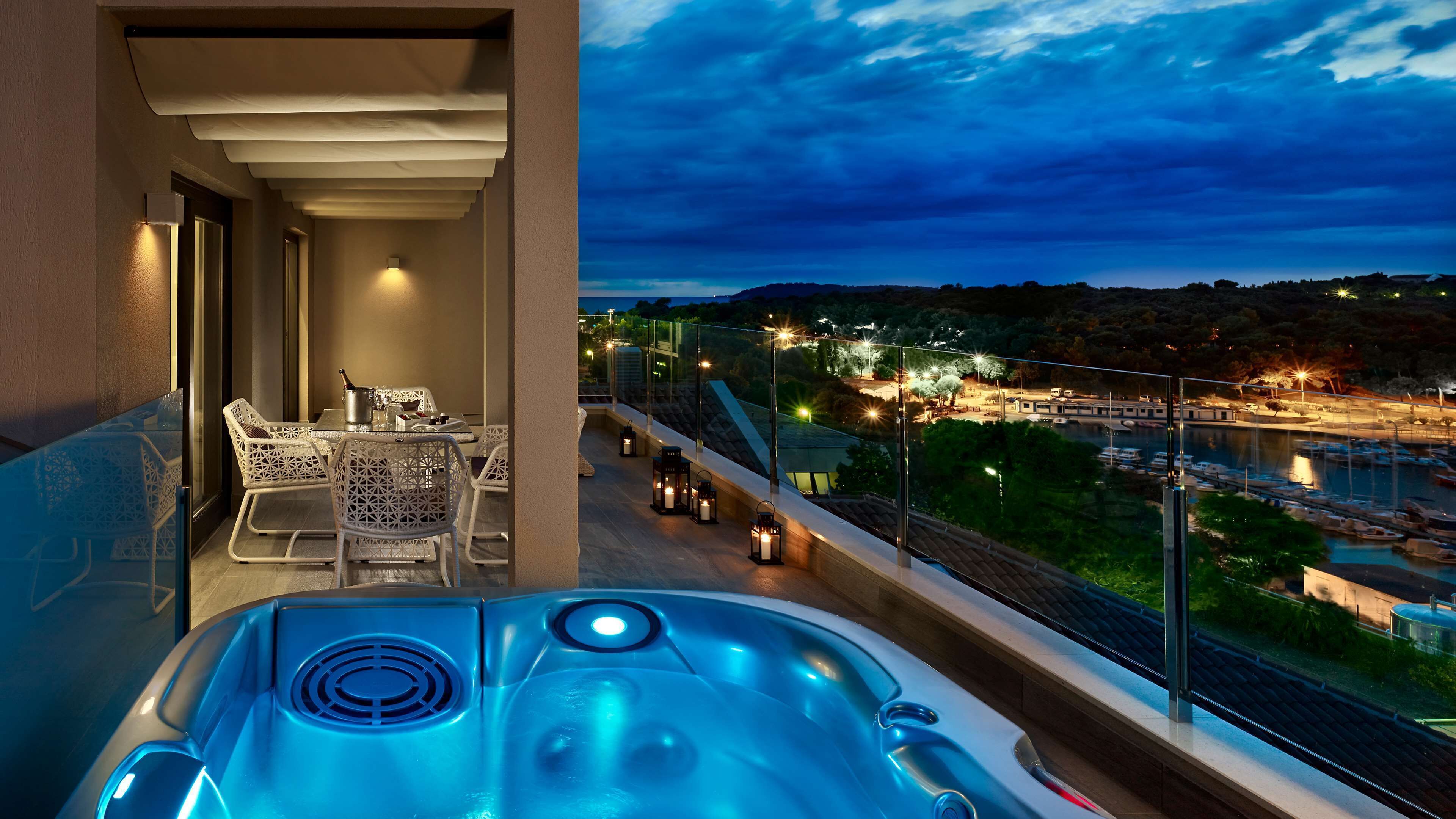 Un balcón con jacuzzi, zona de comedor y vista al atardecer sobre un puerto deportivo.