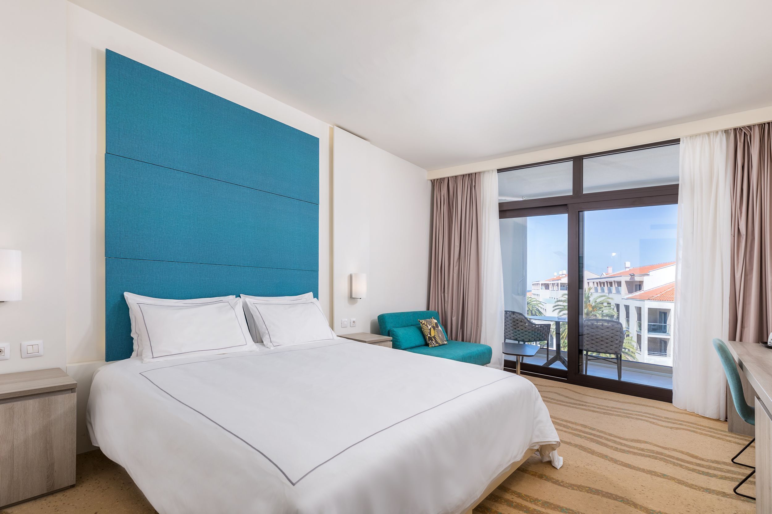 A hotel room with a large bed, a blue accent wall, a balcony with a view, and a small table with chairs.