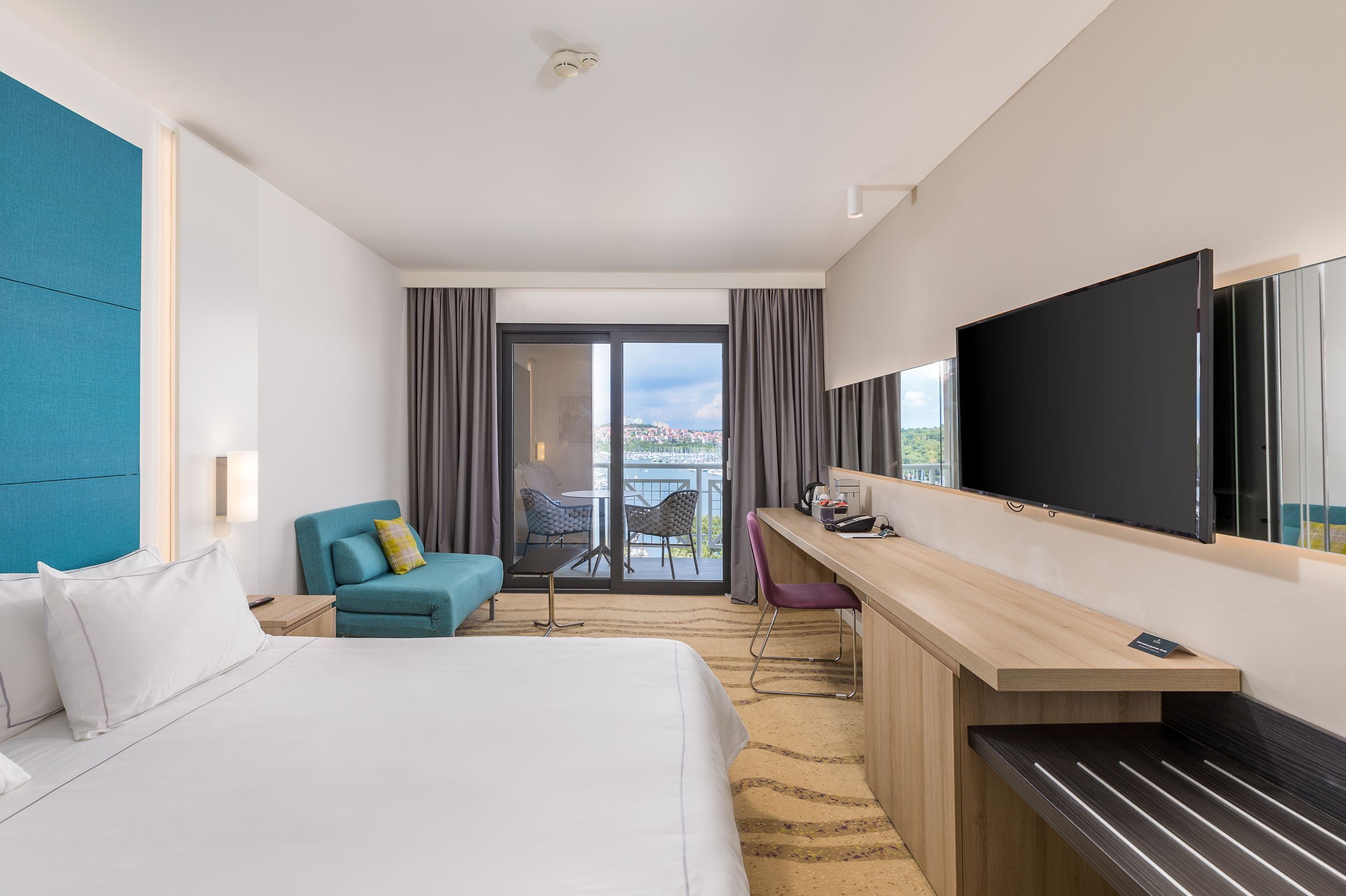 A hotel room with a bed, a desk, a TV, and a balcony with curtains.
