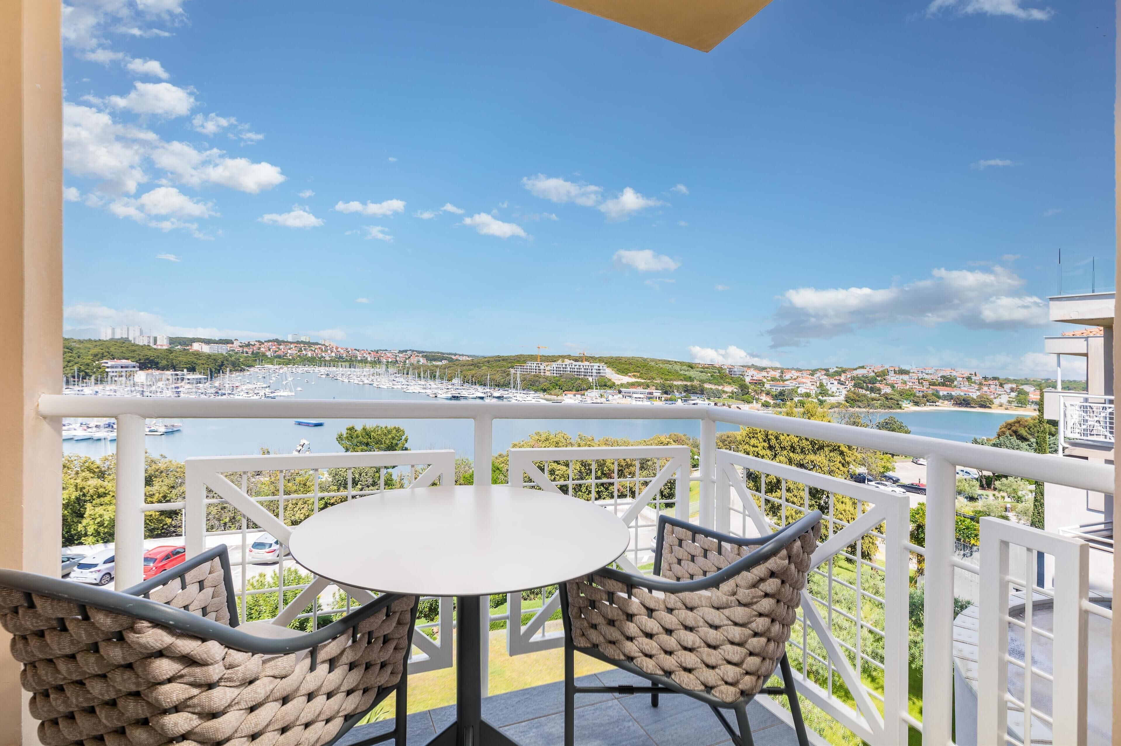 A balcony with a table and chairs, overlooking a marina with boats and a coastal town.