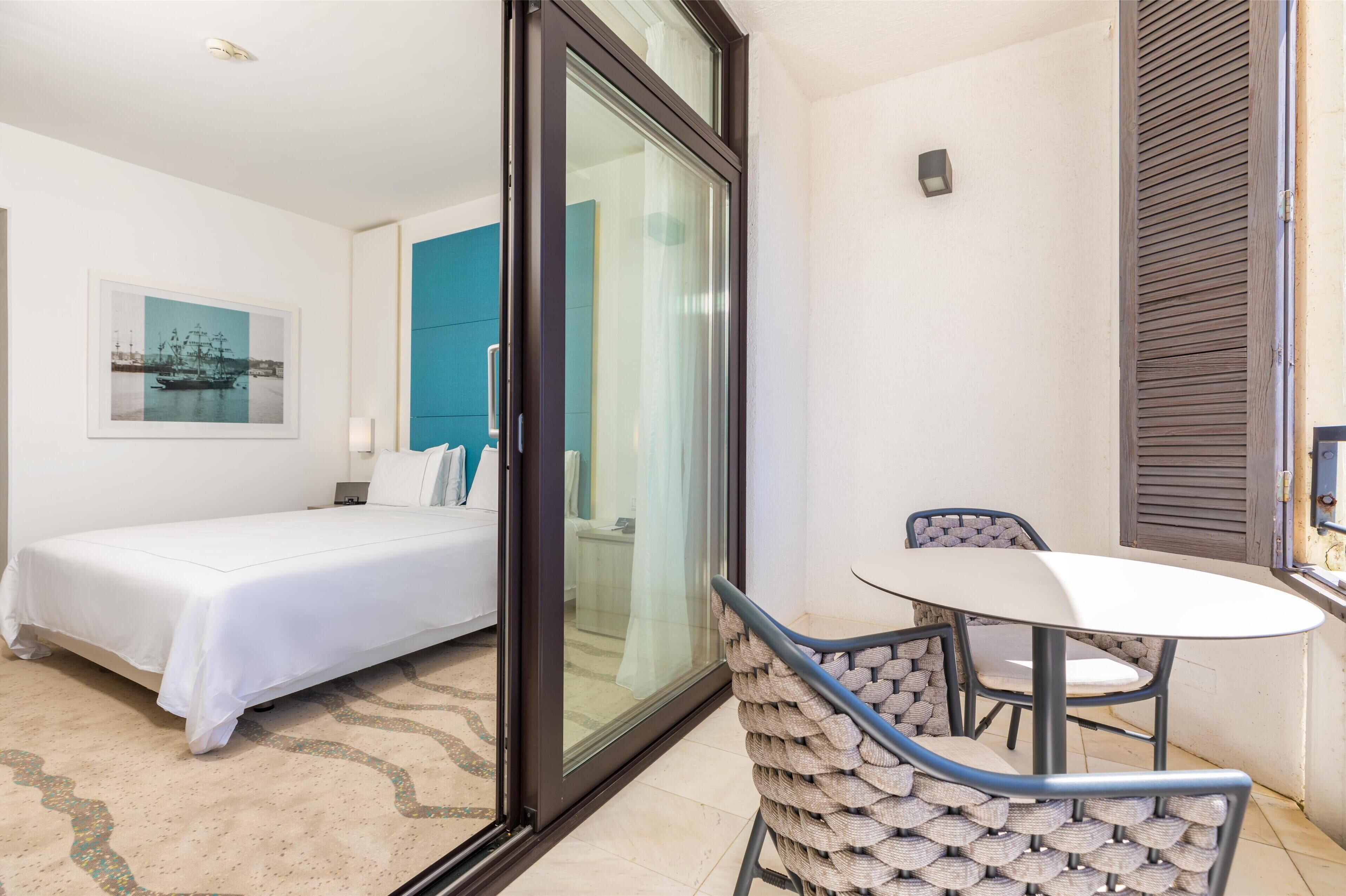 Un dormitorio con una cama, un vidrio que da a un balcón, una mesita y sillas.