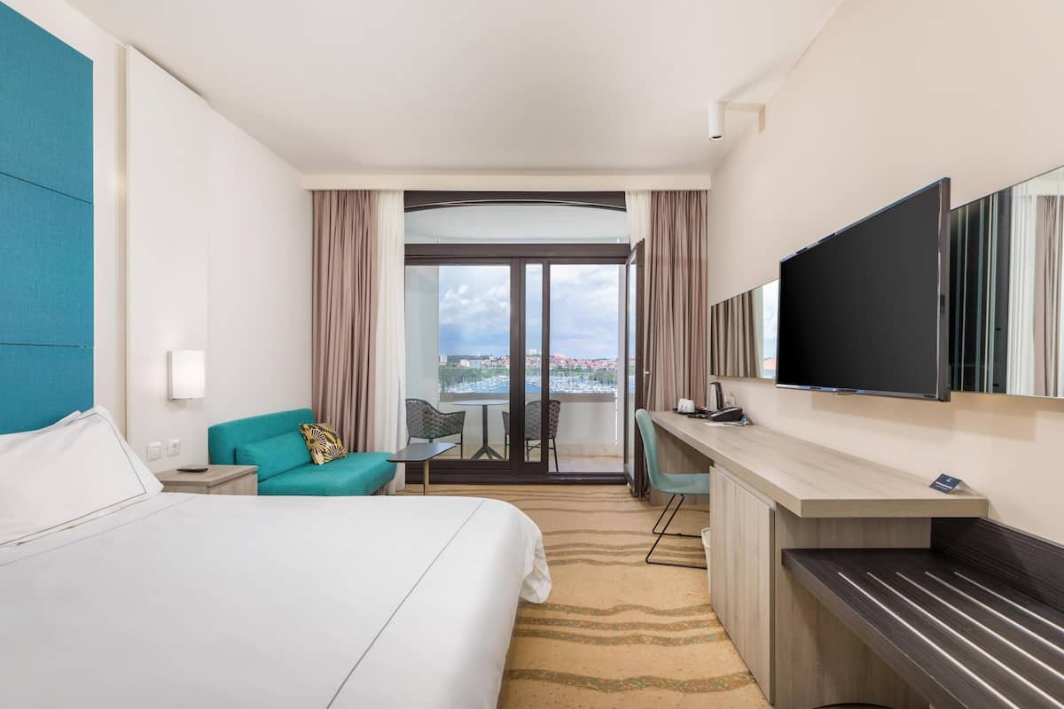 A hotel room with a bed, a desk, a chair, a television, and a view of the city through a large window.