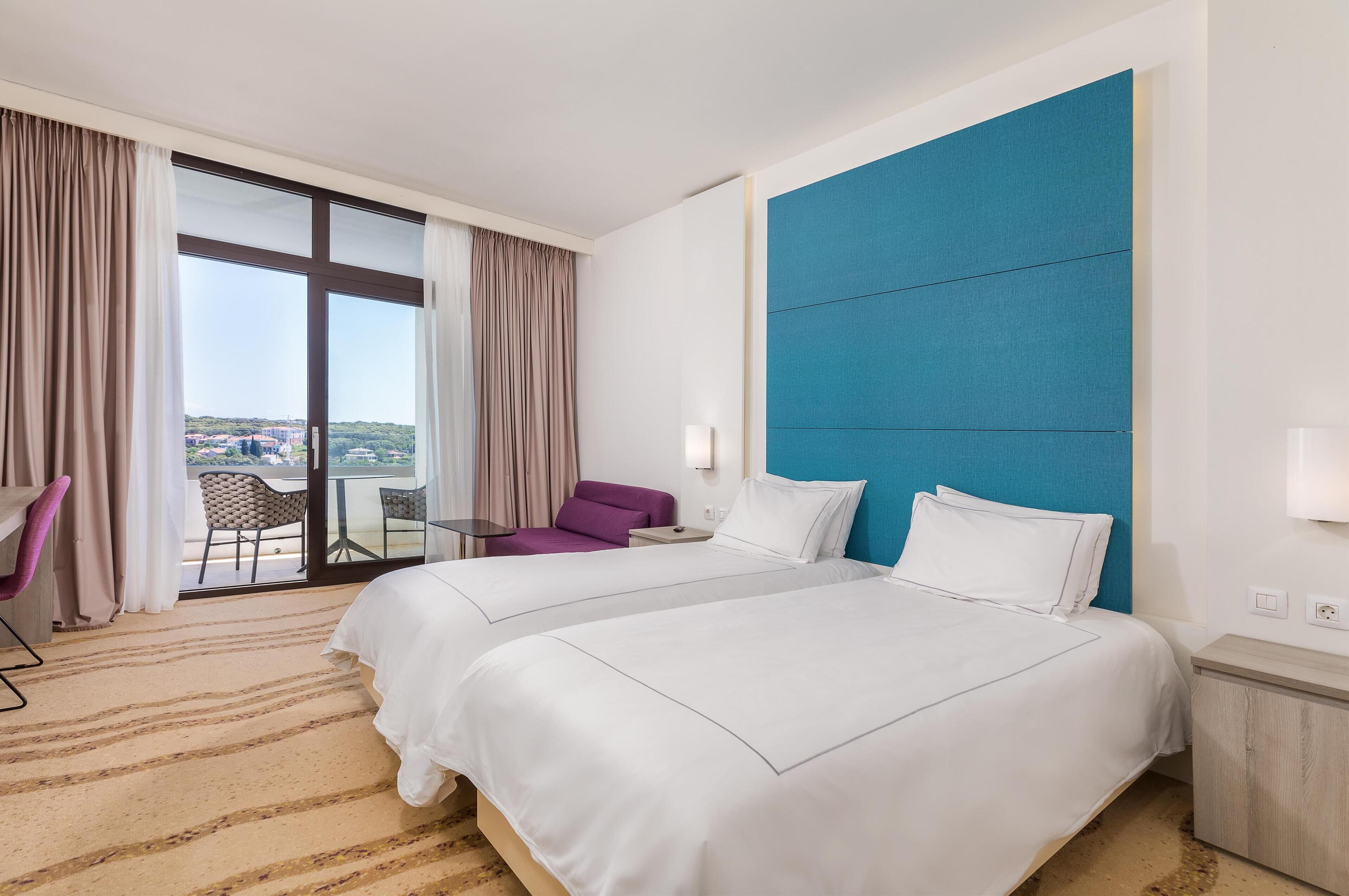 A hotel room with two beds, a large window with curtains, a purple chair, and a view of the city.