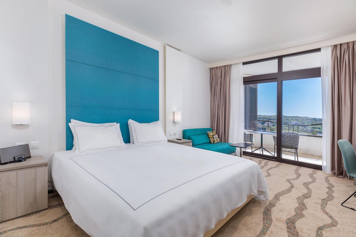 A hotel room with a large bed, a blue accent wall, a balcony with a table and chairs, and a view of the city.
