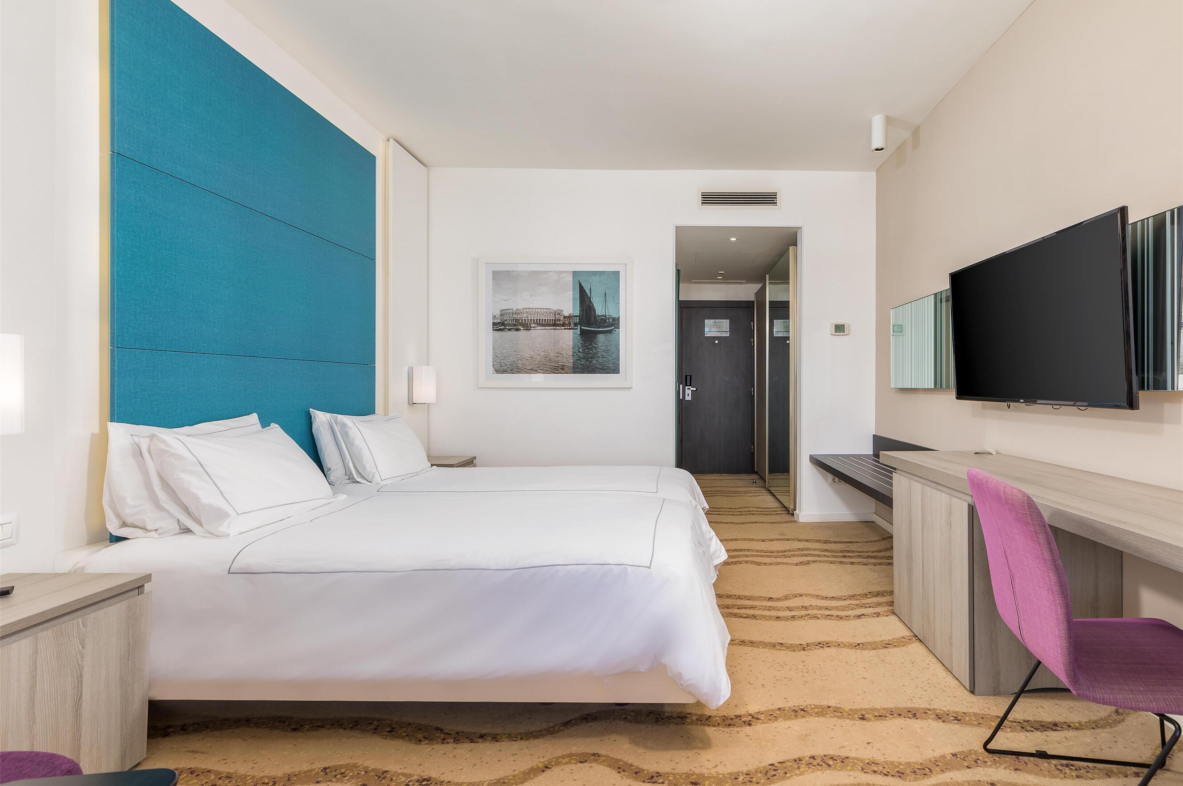 Une chambre d’hôtel avec un grand lit, un bureau avec une chaise, un téléviseur à écran plat et un tableau encadré au mur.