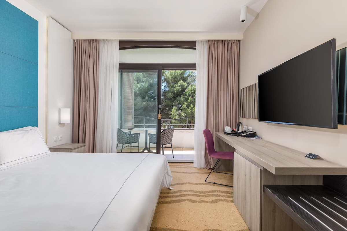 A hotel room with a bed, a desk with a flat-screen TV, a chair, and a balcony with curtains.