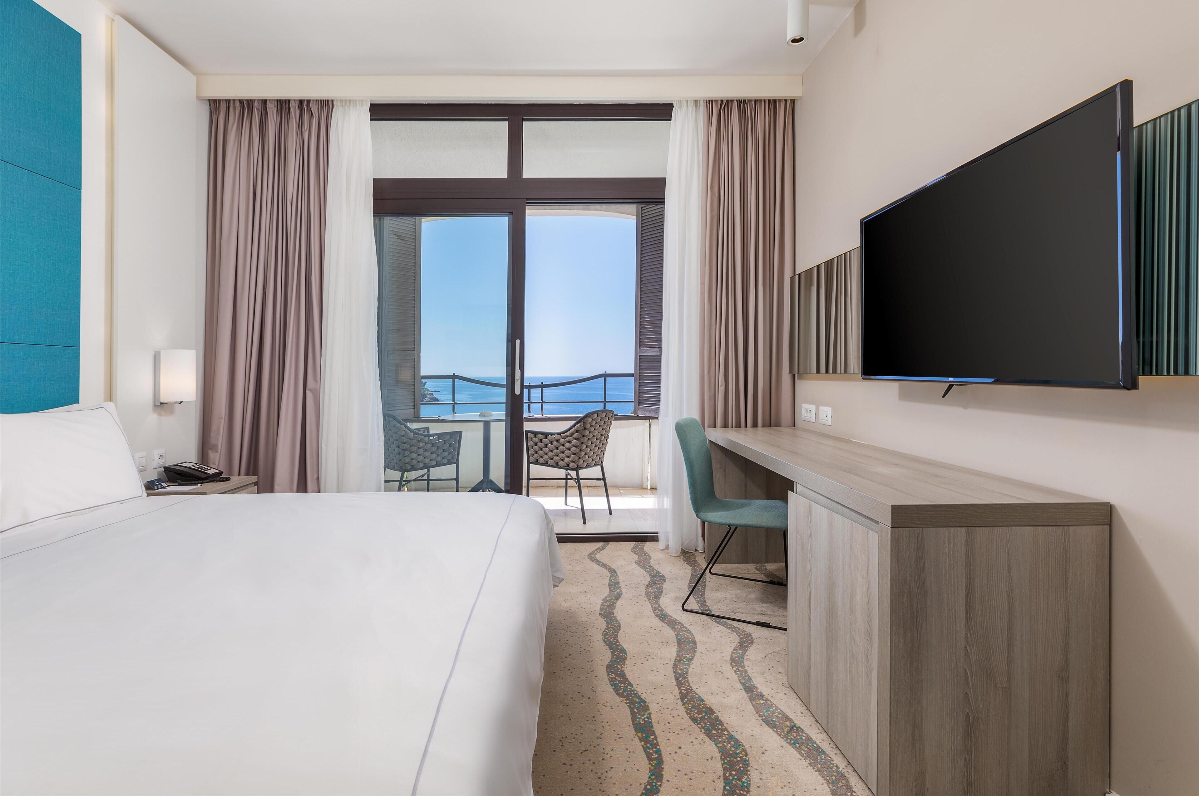Ein Hotelzimmer mit Bett, Schreibtisch, Stuhl, Fernseher und einem Balkon mit Blick.