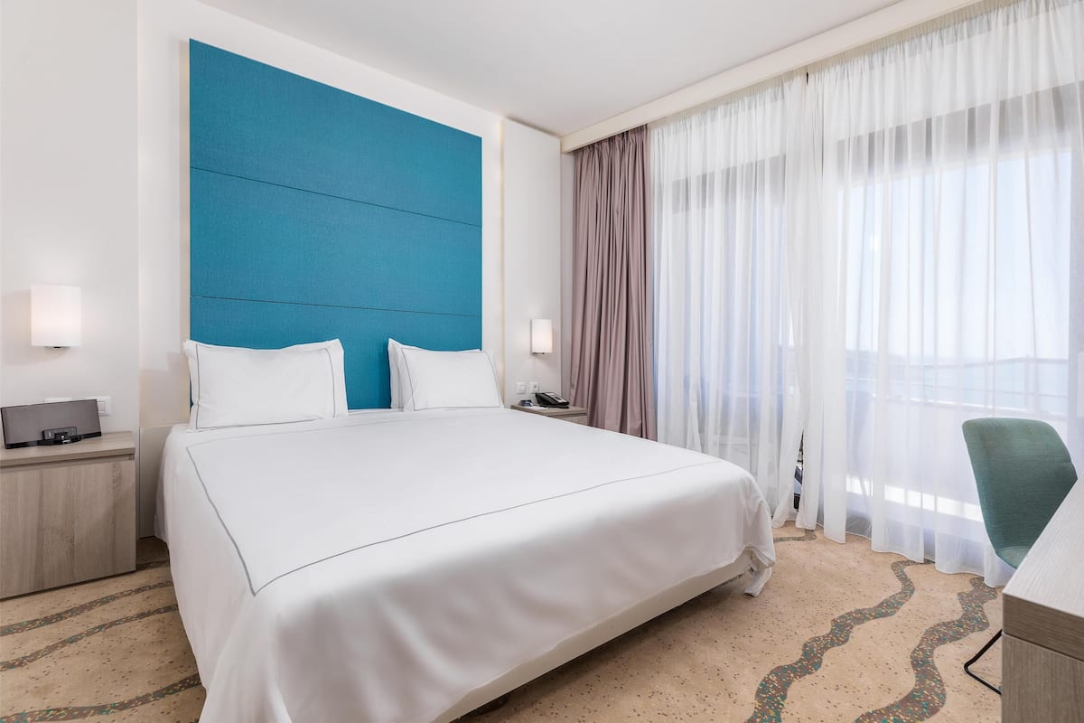 A hotel room with a large bed, a desk, a chair, a nightstand, and a window with sheer curtains.