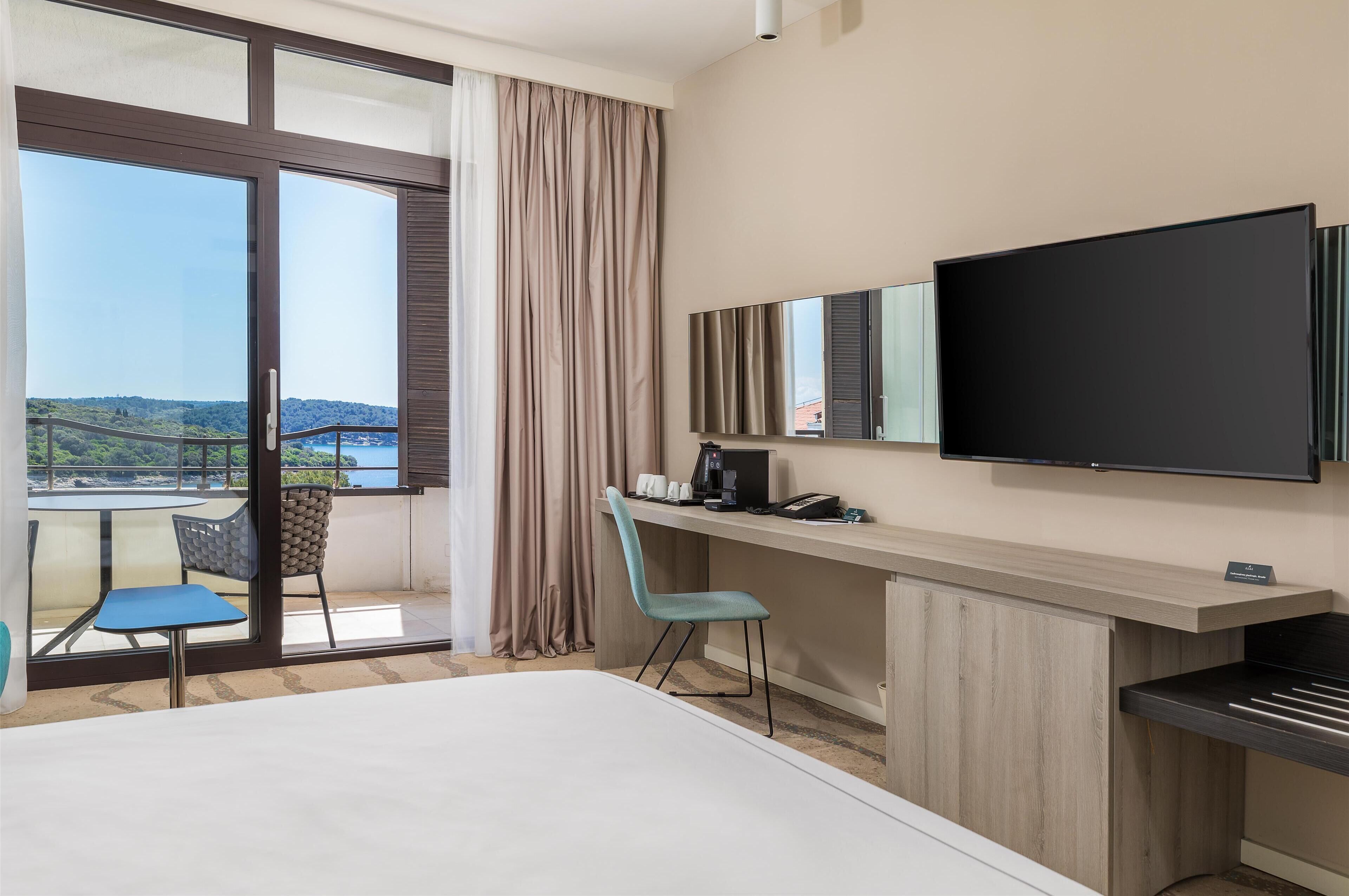 A hotel room with a large bed, a desk with a flat-screen TV, a chair, a small table, and a view of a bridge and greenery through large windows.