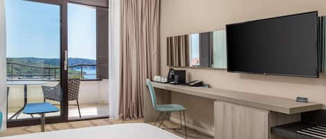 A hotel room with a large bed, a desk with a flat-screen TV, a chair, a small table, and a view of a bridge and greenery through large windows.