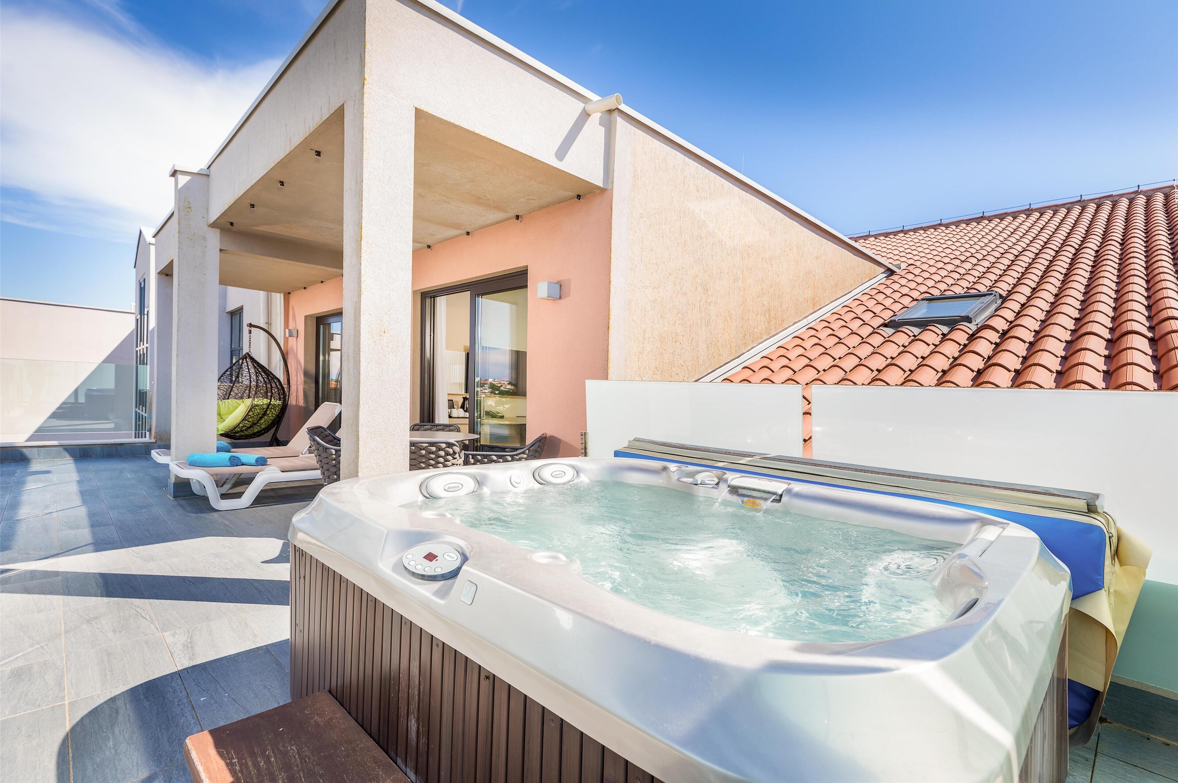 Un jacuzzi sur une terrasse sur le toit, avec un espace salon couvert et une vue sur un bâtiment dont le toit est recouvert de tuiles.
