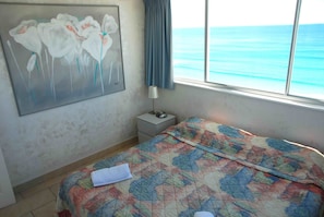 A bedroom with a bed, a nightstand, a lamp, a painting, and a view of the ocean. - The Shore Apartments (Surfers Paradise)