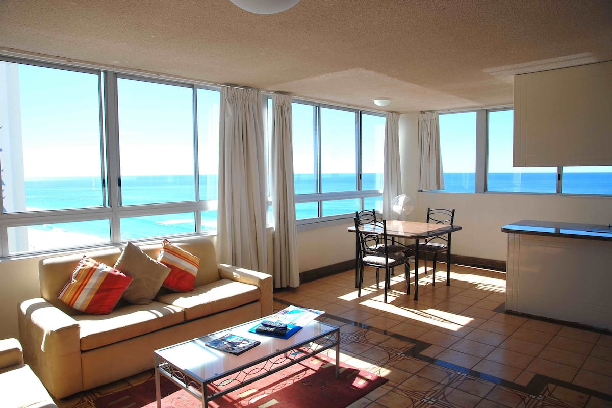 A living room with a sofa, coffee table, and dining table with chairs. There are large windows with curtains, a view of the ocean, and a kitchen area with a counter.