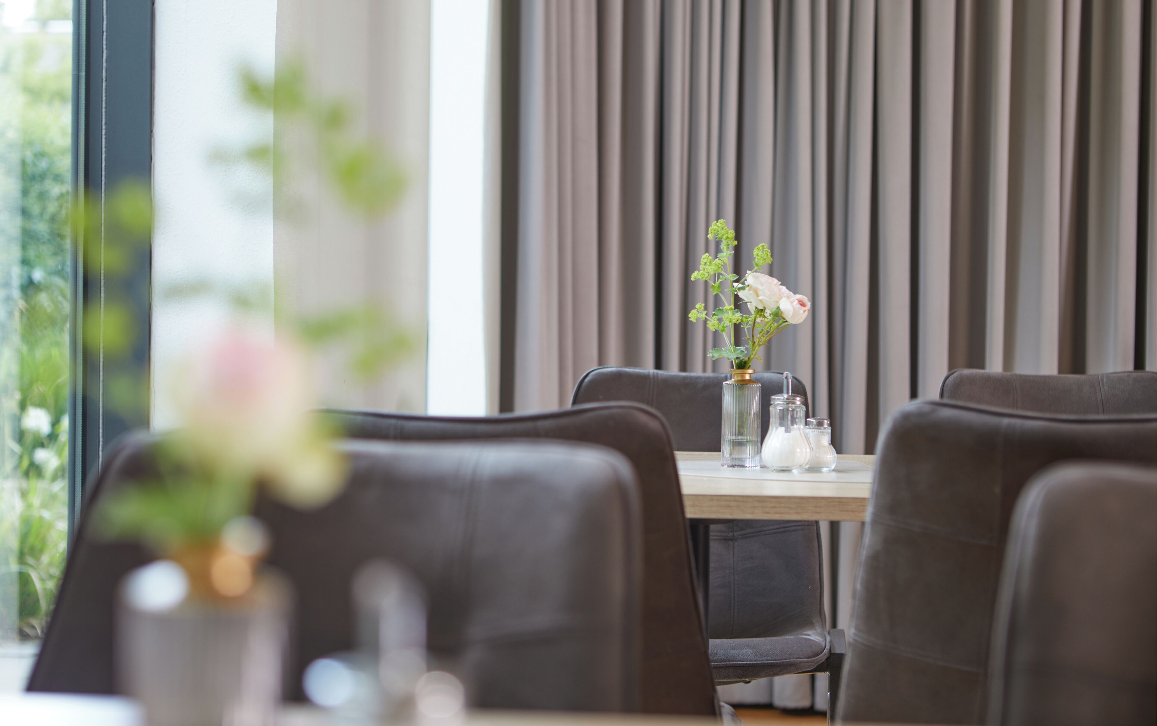 Un coin repas avec une table dressée pour deux personnes, agrémenté d’un vase avec des fleurs, d’un salier et d’un poivrier, et d’un verre.