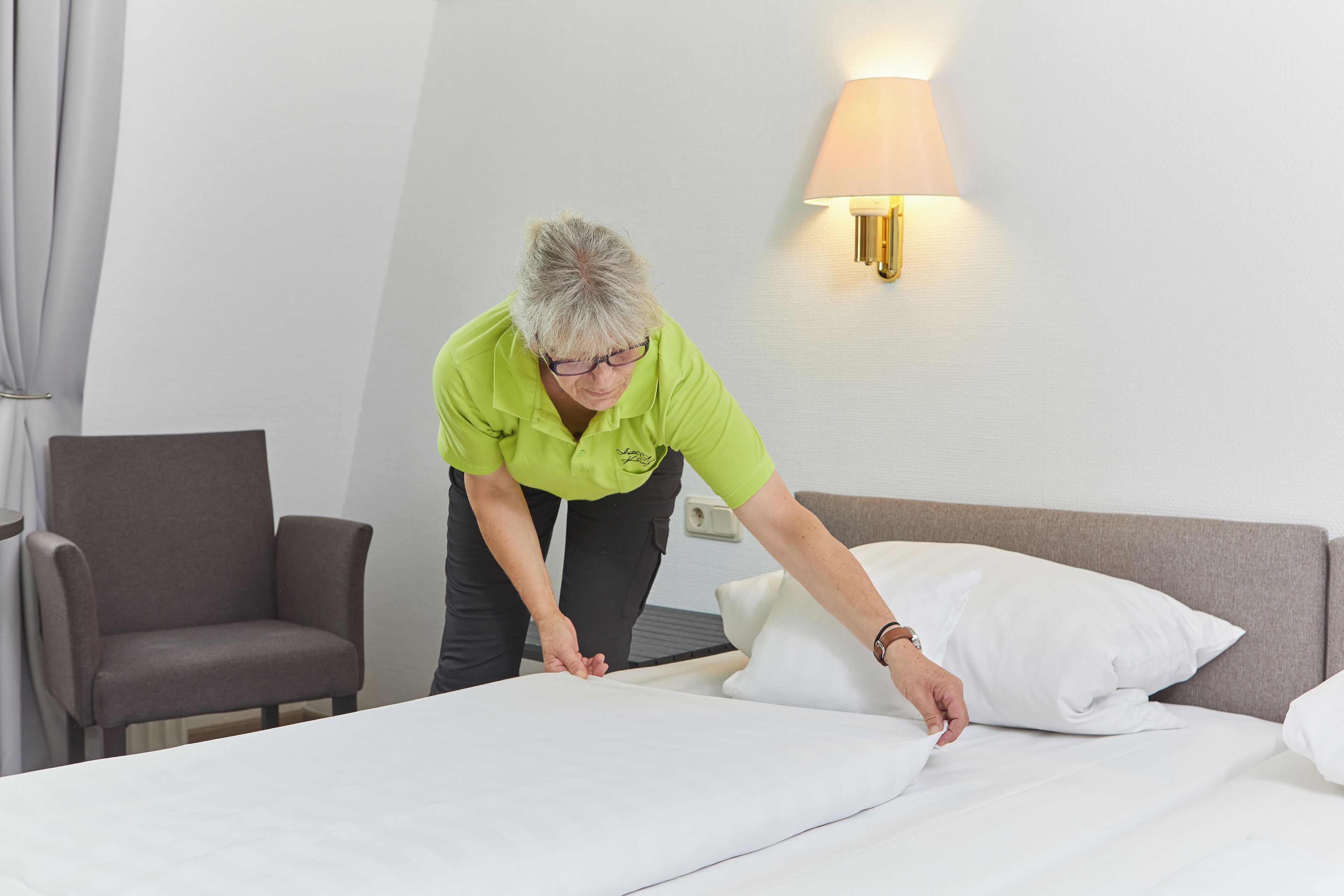 A hotel room with a bed, a chair, and a wall-mounted lamp.
