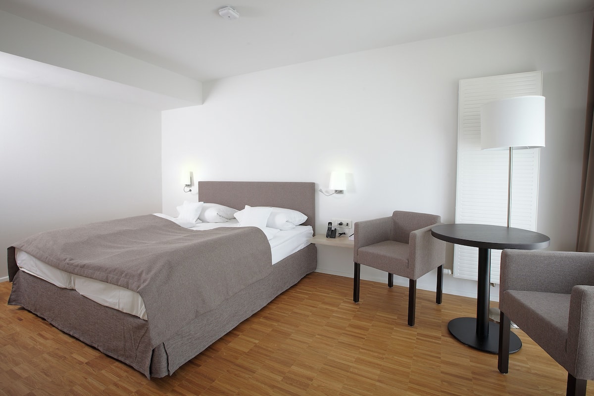 Ein modernes Hotelzimmer mit einem aufgeräumten Bett, einem runden Tisch, zwei Sesseln und einer Stehlampe.