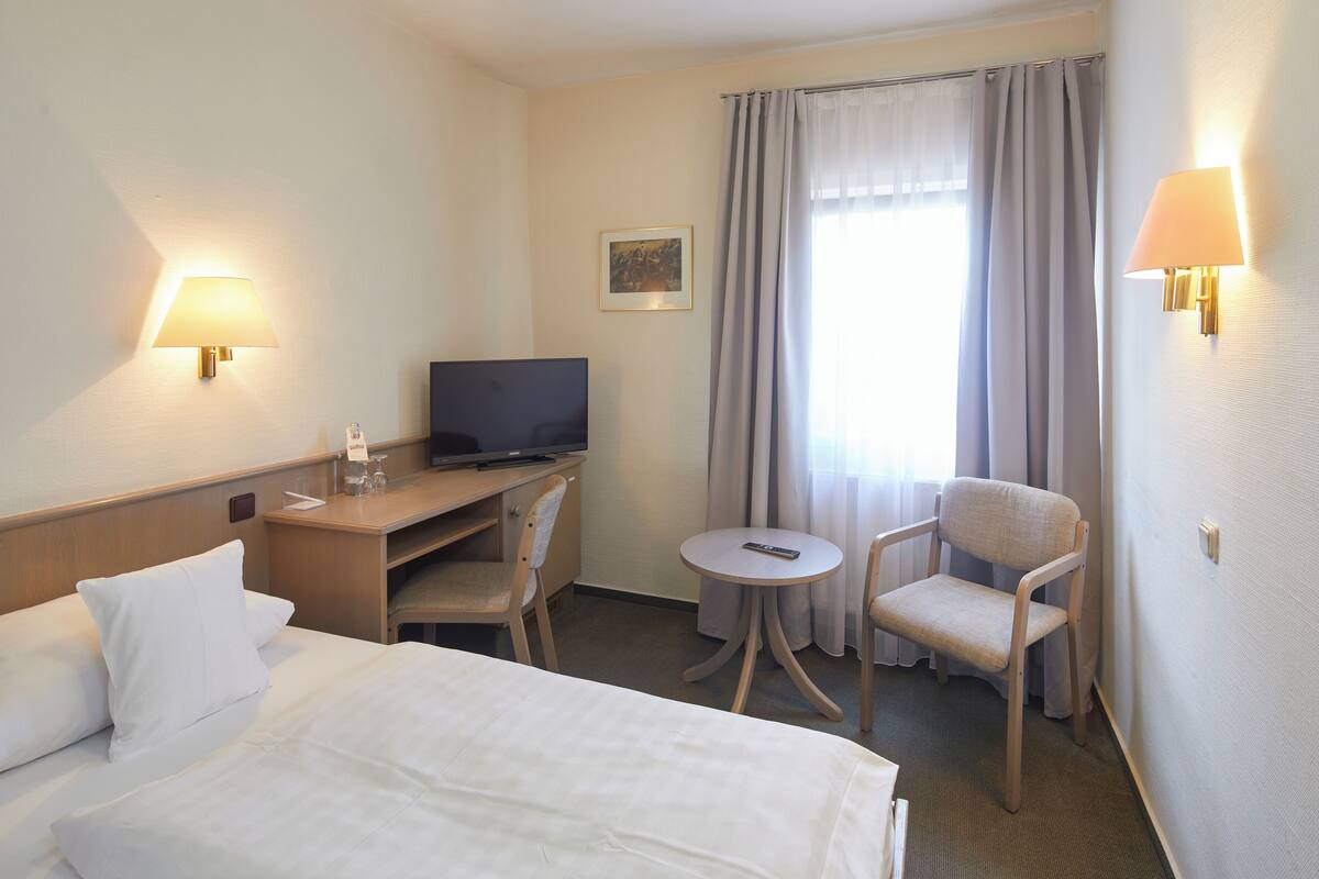 Ein Hotelzimmer mit einem Bett, einem Schreibtisch mit Fernseher, einem Sessel, einem kleinen Tisch und zwei Wandlampen.