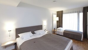 Ein Hotelzimmer mit einem großen Bett, einem kleineren Bett, einem Nachttisch, einer Lampe und einem Fenster mit Vorhängen.