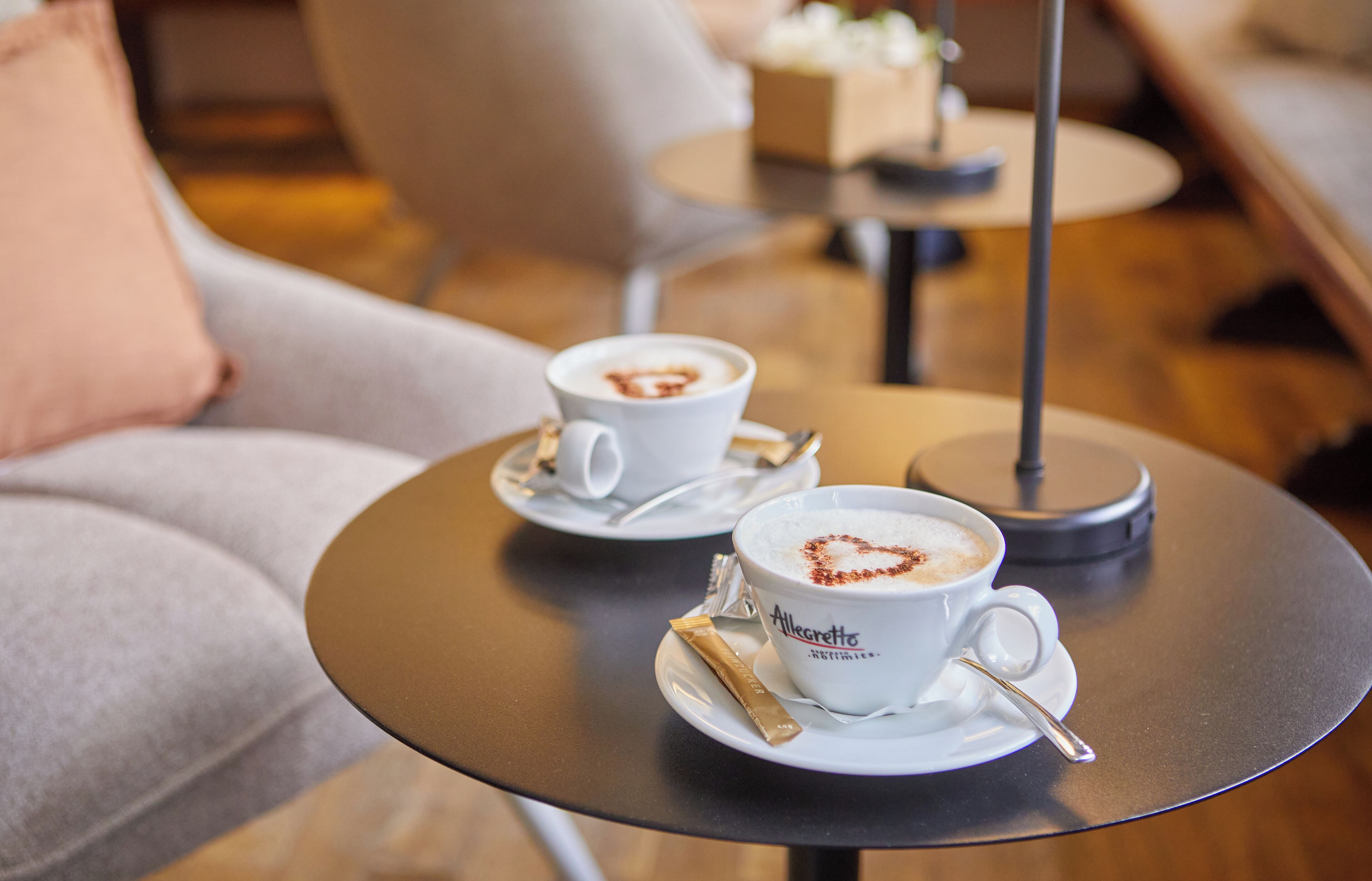 Two white cups of coffee on a round table with a heart design on one cup, a small box, and a chair in the background.