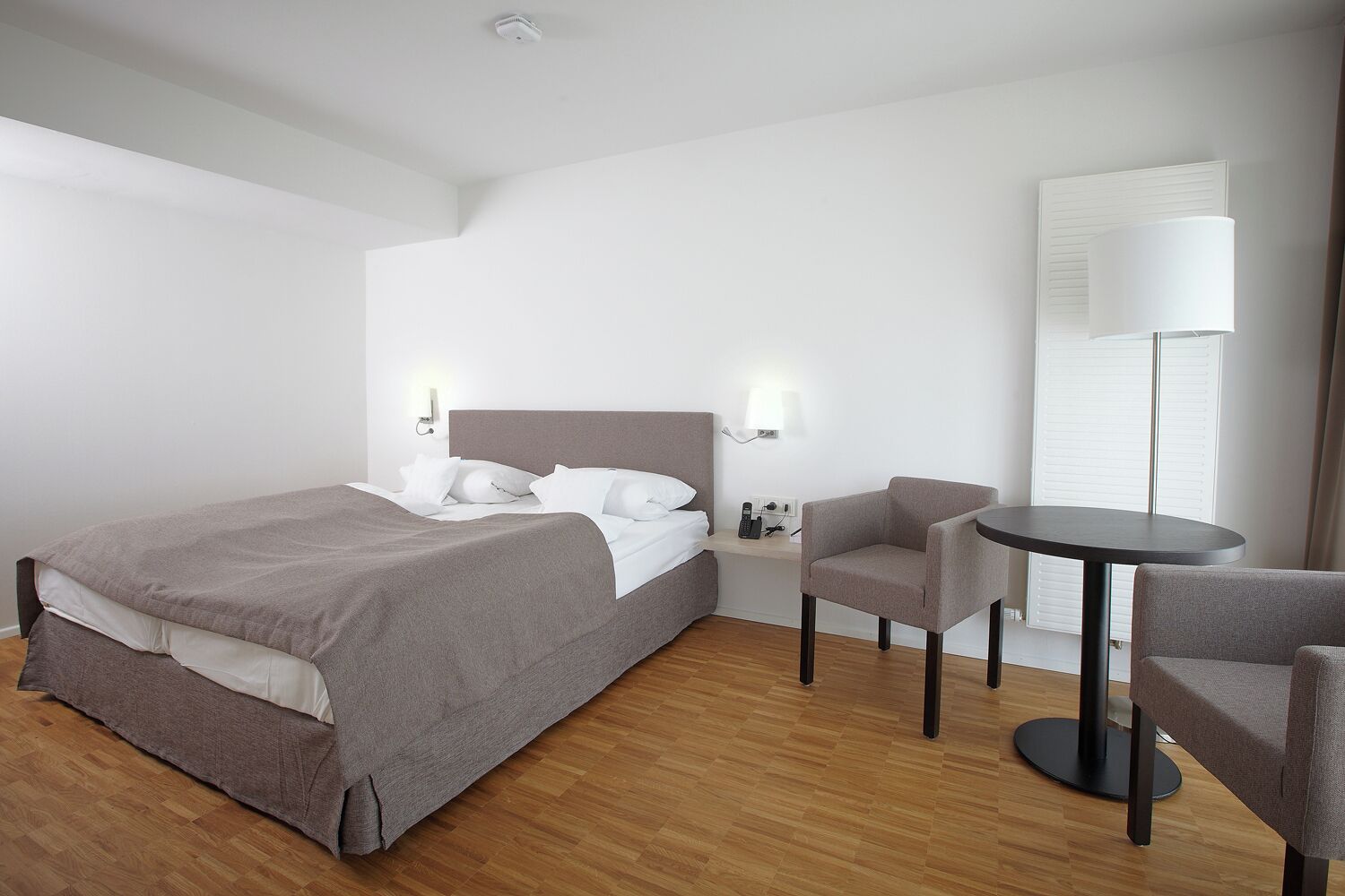 Una habitación de hotel con una cama, una silla, una mesita pequeña y una mesita de noche con lámpara.