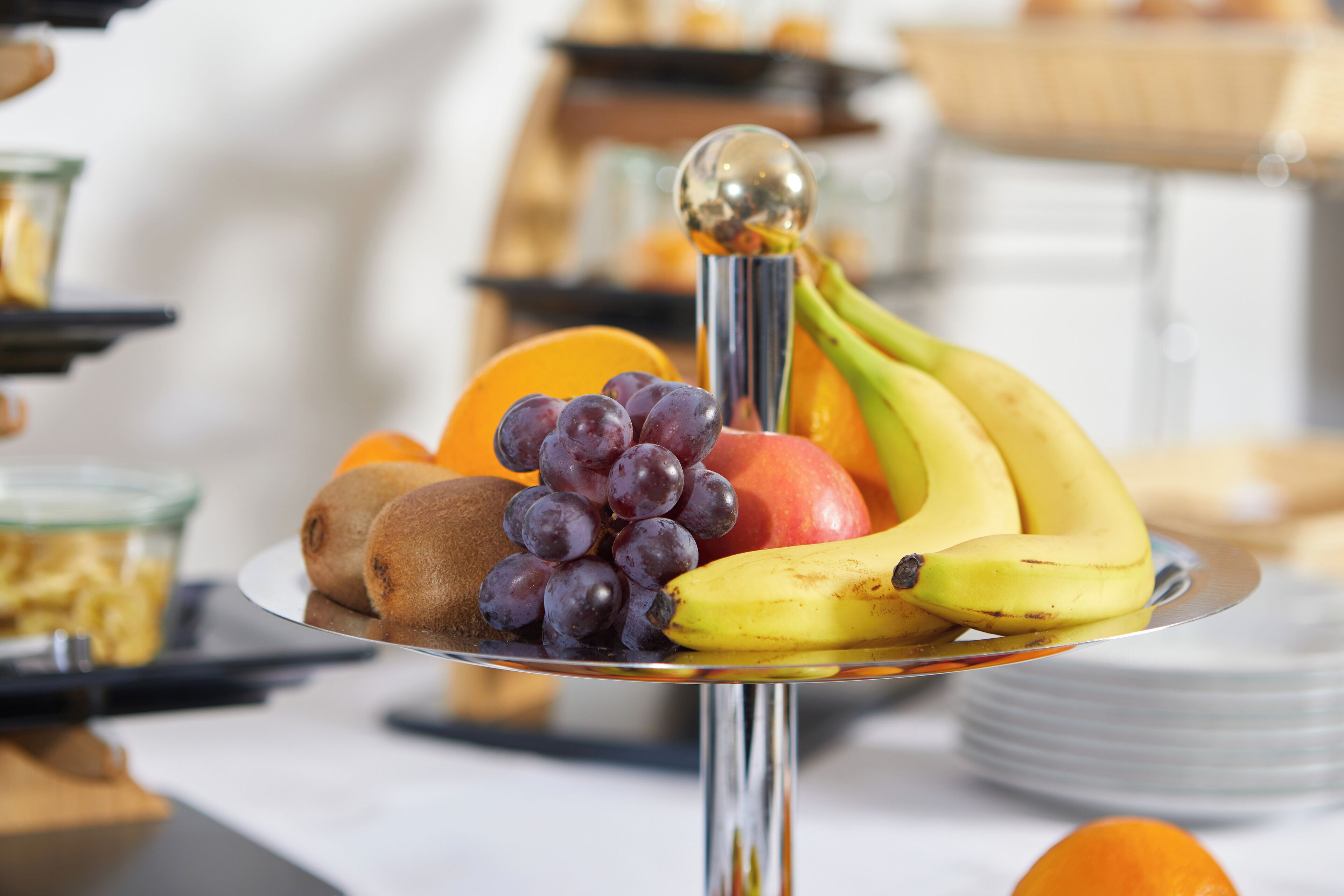 Una bandeja de frutas con plátanos, uvas, kiwi y rodajas de naranja sobre un soporte metálico reflectante.