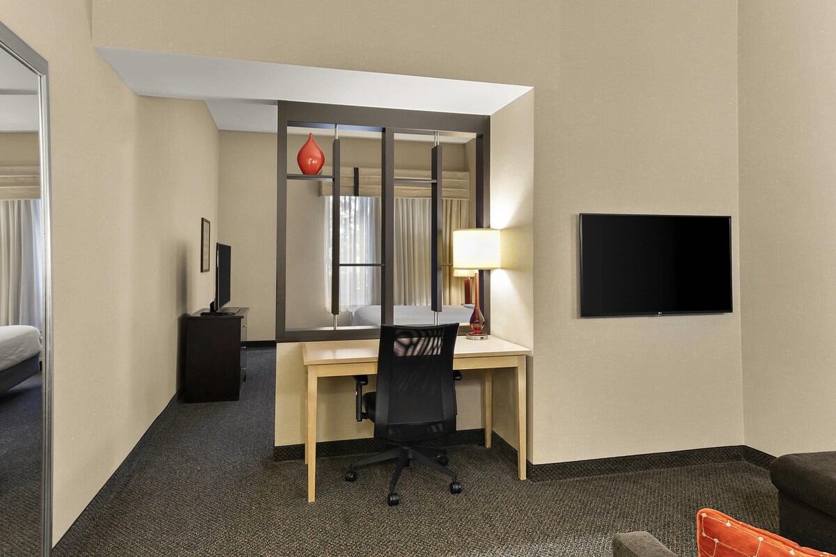 A hotel room with a desk, chair, bed, TV, and a mirror.